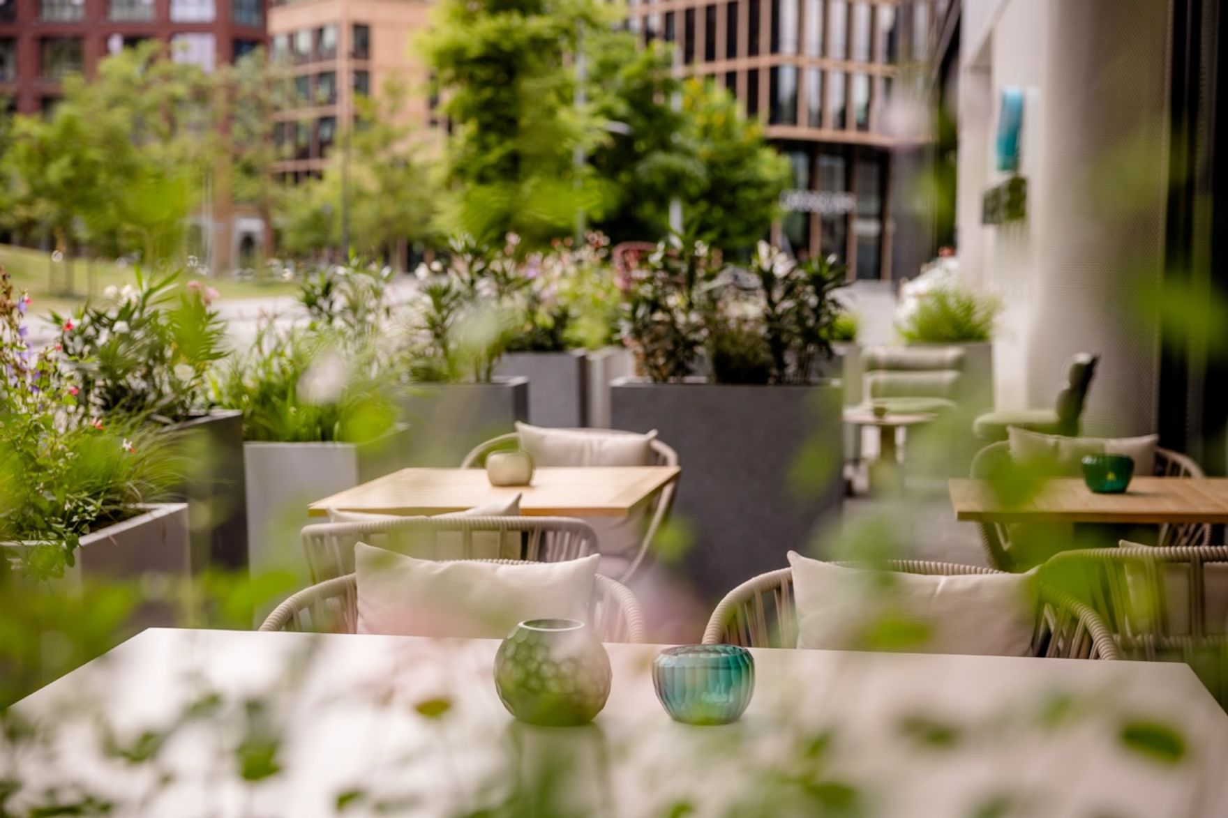 Terrassenbereich eines Hotels mit gemütlichen Sitzplätzen, weißen Tischen, bunten Windlichtern und Blick auf grüne Pflanzen sowie moderne Stadtgebäude im Hintergrund.