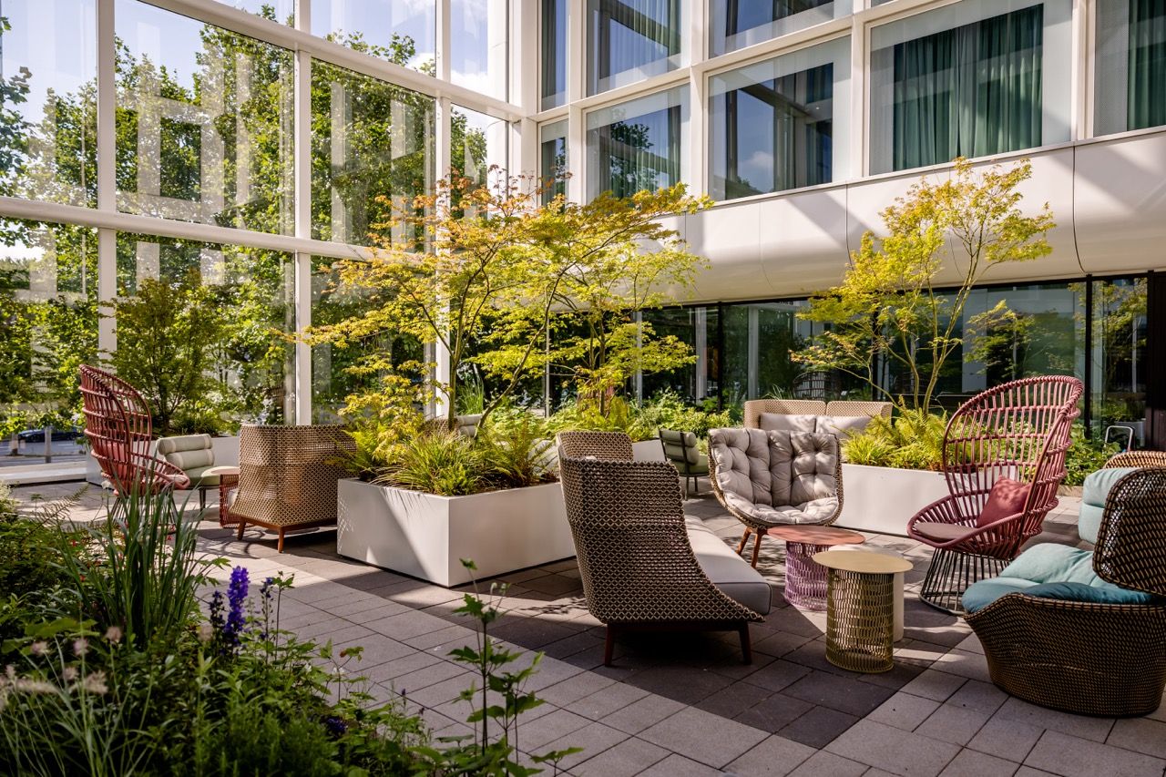 Helle, moderne Innenhof-Lounge mit bequemen Sitzmöbeln und üppiger Begrünung, umgeben von großen Glasfenstern, die viel Tageslicht hereinlassen. Ruhiger Ort zum Entspannen und Genießen der Natur im urbanen Ambiente.