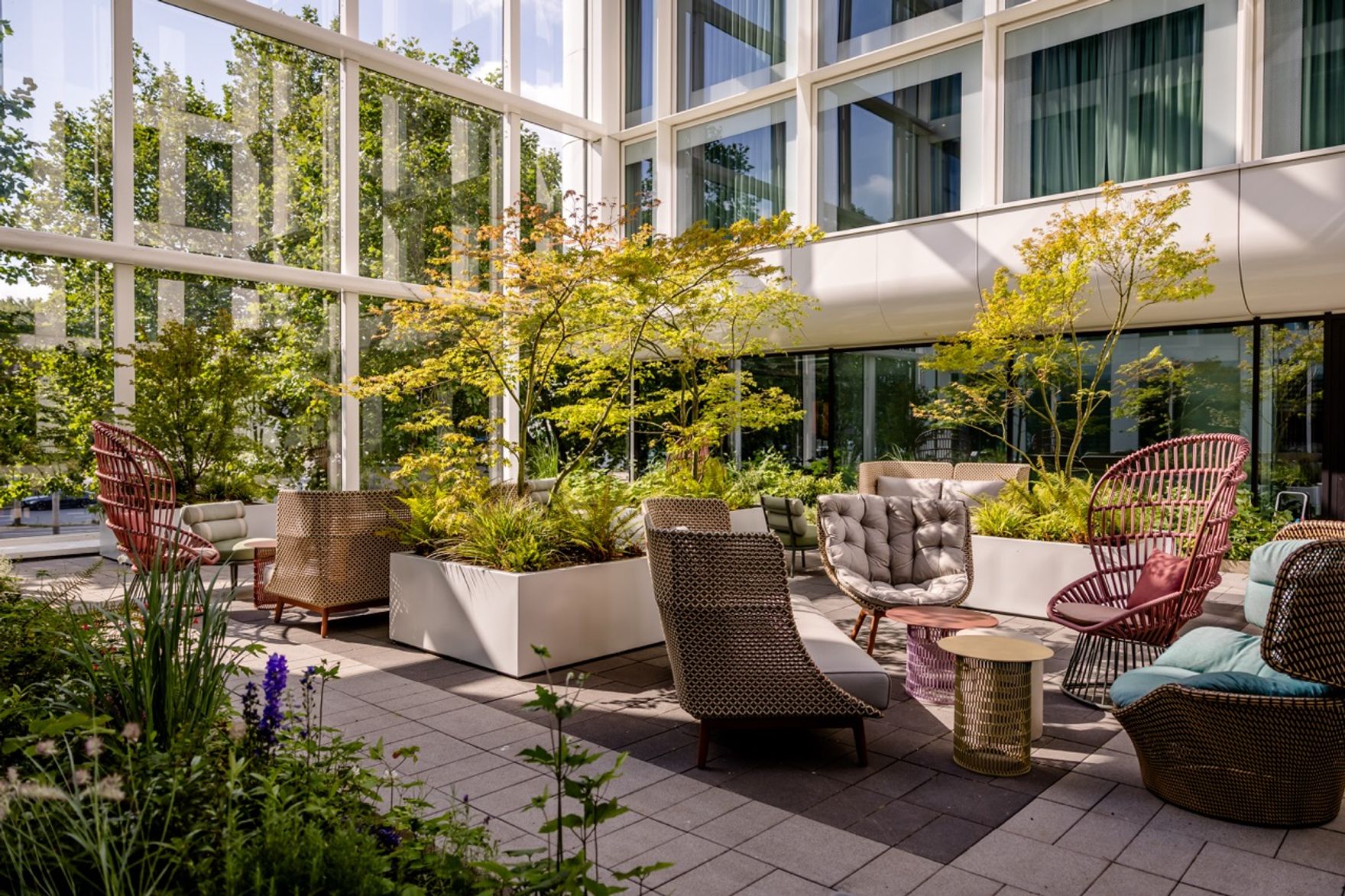 Helle, moderne Innenhof-Lounge mit bequemen Sitzmöbeln und üppiger Begrünung, umgeben von großen Glasfenstern, die viel Tageslicht hereinlassen. Ruhiger Ort zum Entspannen und Genießen der Natur im urbanen Ambiente.