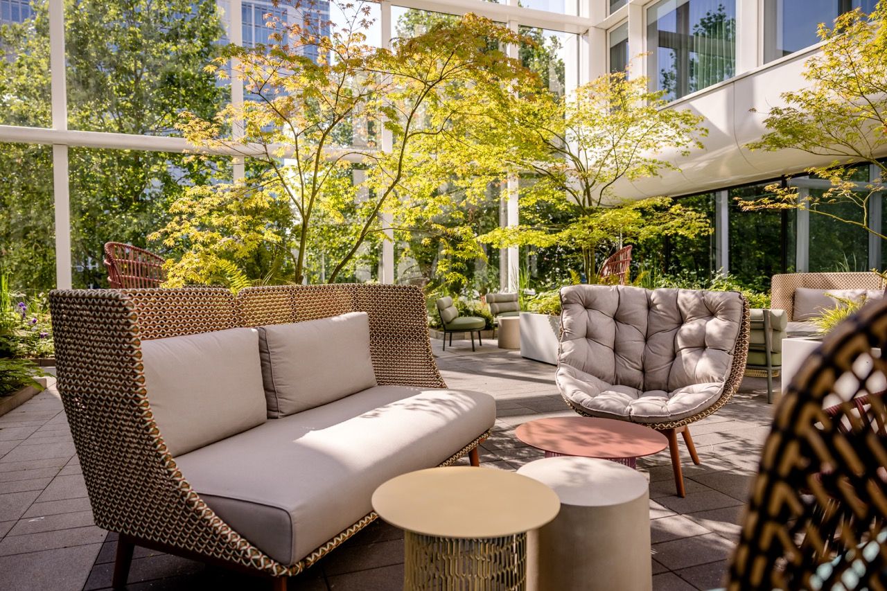 Helle, moderne Lounge im Wintergarten mit gemütlichen Sesseln und Sofas, umgeben von grün belaubten Bäumen, ideal für entspannte Gespräche und Pausen in natürlicher Atmosphäre.