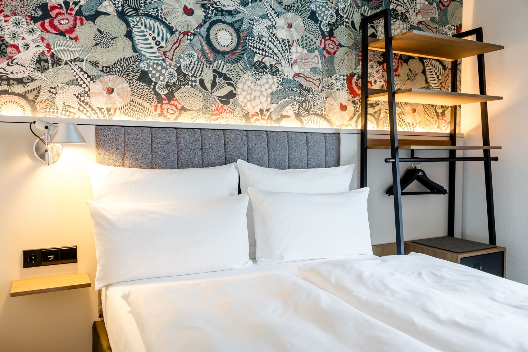 Modernes Hotelzimmer mit großem, weiß bezogenem Doppelbett, grauem gepolstertem Kopfteil und dekorativer Wand mit floralem Muster. Helle Beleuchtung sorgt für freundliche Atmosphäre. Offenes Regalsystem bietet Ablage- und Aufhängemöglichkeiten.