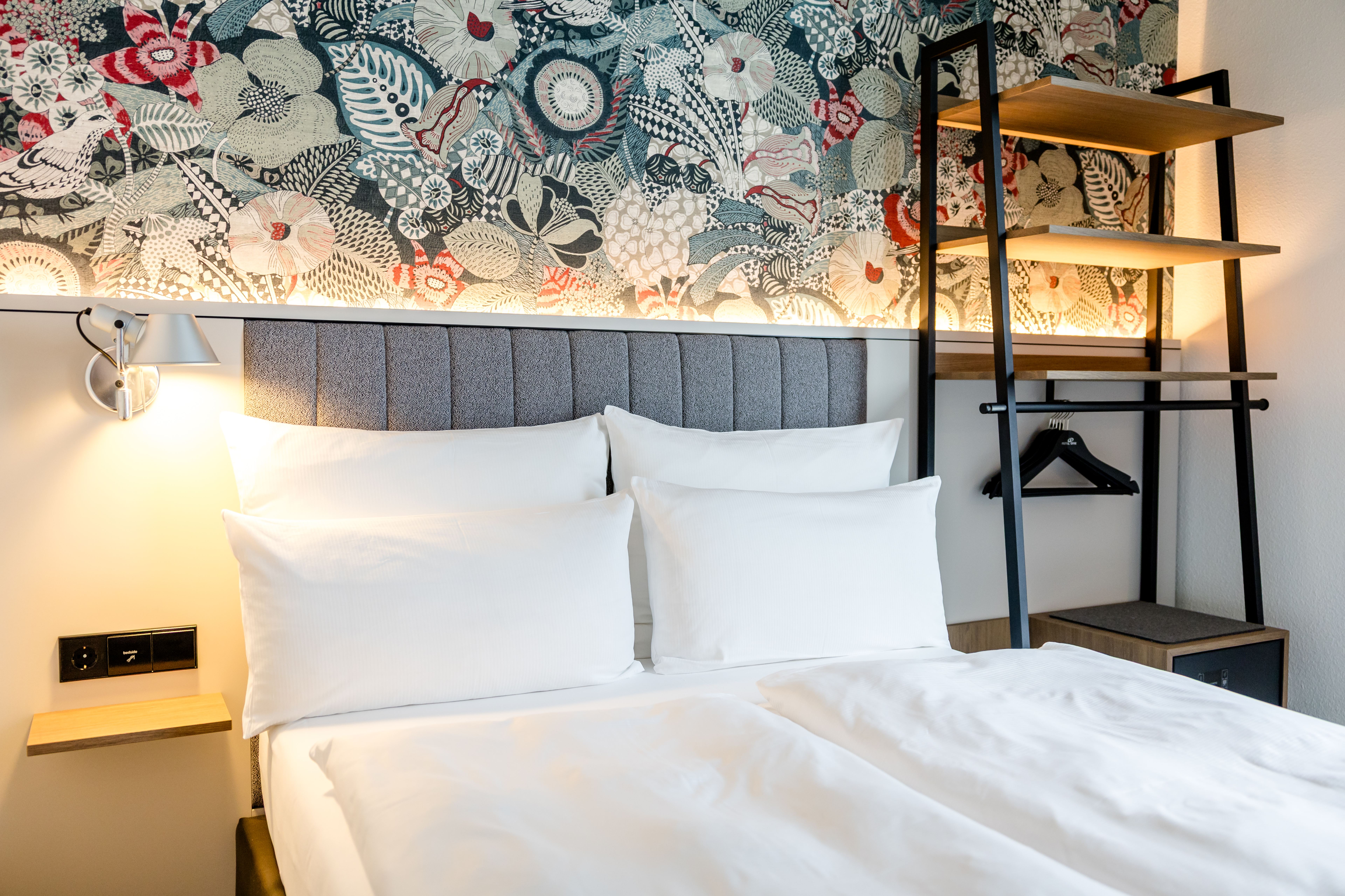 Modernes Hotelzimmer mit großem, weiß bezogenem Doppelbett, grauem gepolstertem Kopfteil und dekorativer Wand mit floralem Muster. Helle Beleuchtung sorgt für freundliche Atmosphäre. Offenes Regalsystem bietet Ablage- und Aufhängemöglichkeiten. Modernes Hotelzimmer mit großem, weiß bezogenem Doppelbett, grauem gepolstertem Kopfteil und dekorativer Wand mit floralem Muster. Helle Beleuchtung sorgt für freundliche Atmosphäre. Offenes Regalsystem bietet Ablage- und Aufhängemöglichkeiten.