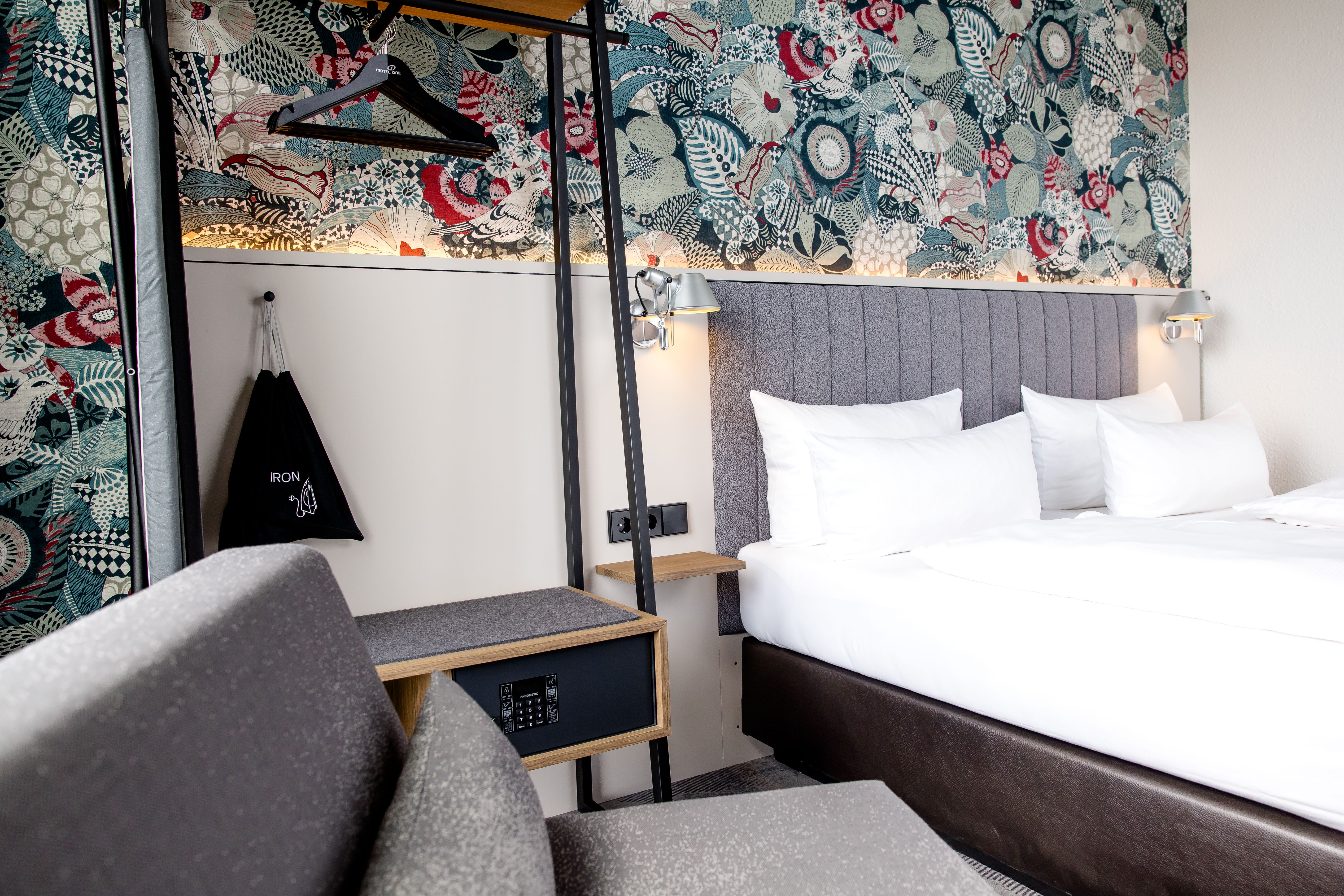 Modern eingerichtetes Hotelzimmer mit großzügigem Doppelbett, gemütlicher Sitzecke, elegantem Muster an der Wand und funktionalem Kleiderschrank mit Spiegel und Safe für Komfort und Sicherheit während des Aufenthalts.