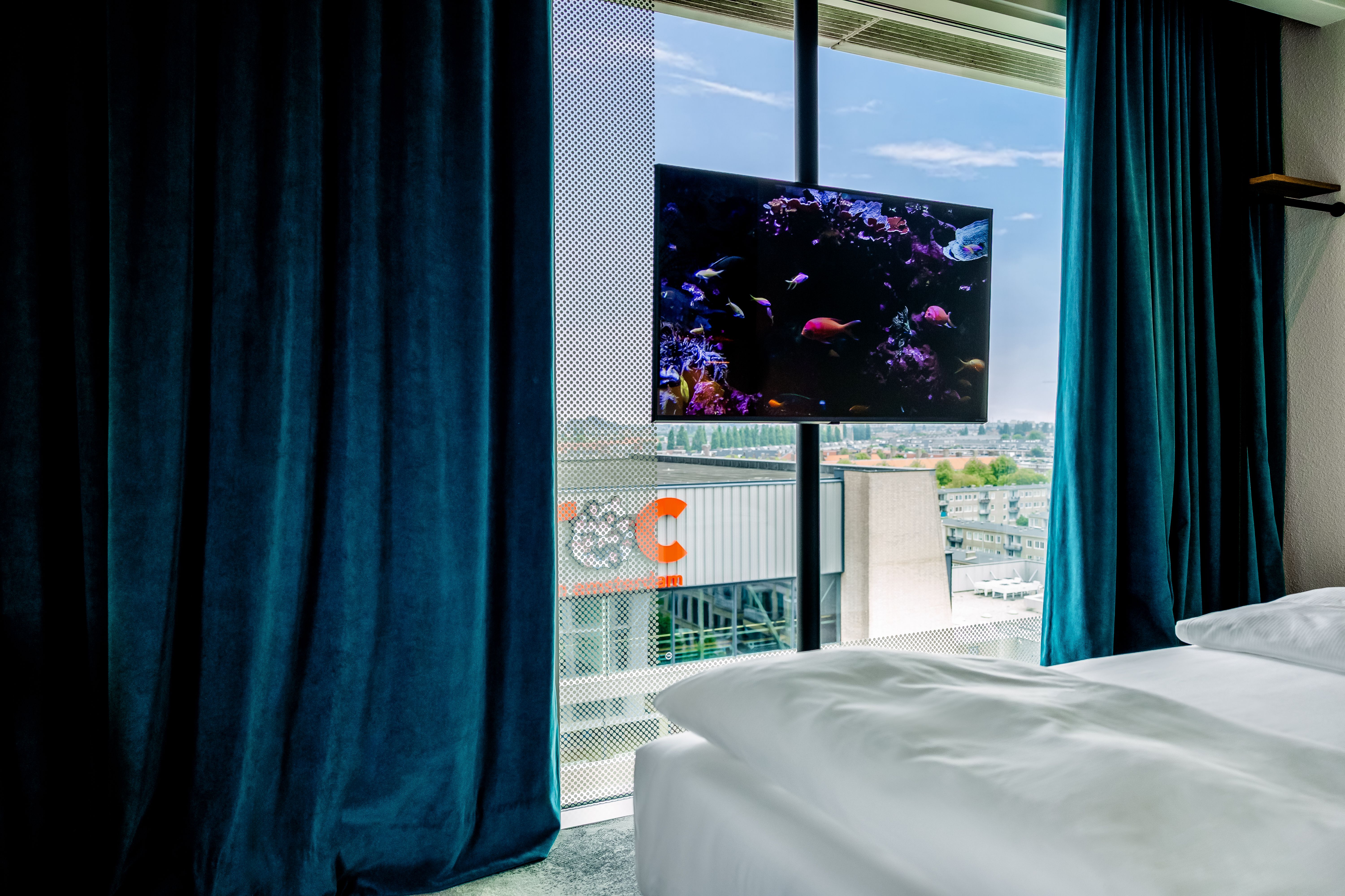 Schlafzimmer mit großen Fenstern, dunkelblauen Vorhängen und Blick auf Stadt. Fernseher mit Aquarium-Motiv hängt vor dem Fenster, gemütliche und helle Atmosphäre.