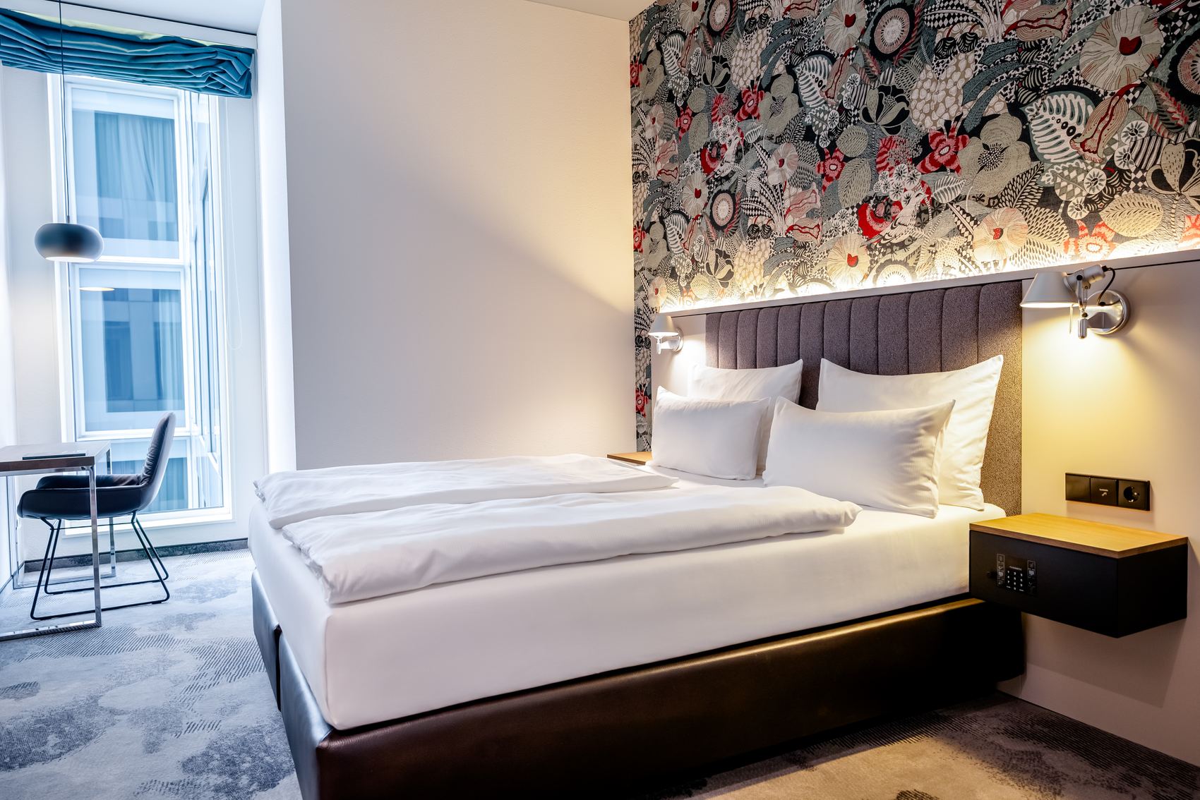 Modern eingerichtetes Hotelzimmer mit großem, weiß bezogenem Doppelbett, dekorativer Tapete hinter dem Kopfteil und Arbeitsplatz am Fenster mit Stuhl. Helle, freundliche Atmosphäre und indirekte Beleuchtung sorgen für Wohlgefühl.