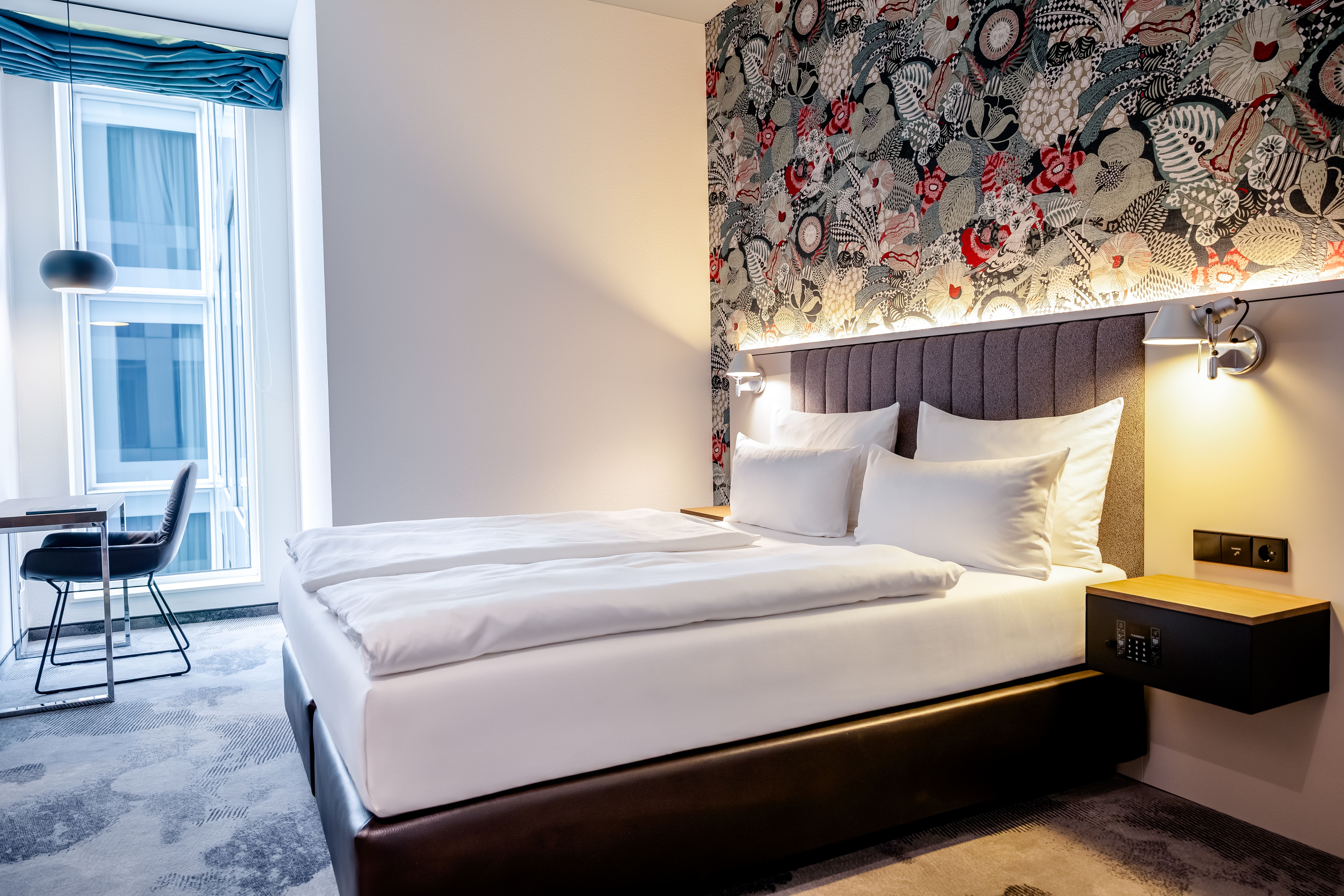 Modern eingerichtetes Hotelzimmer mit großem, weiß bezogenem Doppelbett, dekorativer Tapete hinter dem Kopfteil und Arbeitsplatz am Fenster mit Stuhl. Helle, freundliche Atmosphäre und indirekte Beleuchtung sorgen für Wohlgefühl. Modern eingerichtetes Hotelzimmer mit großem, weiß bezogenem Doppelbett, dekorativer Tapete hinter dem Kopfteil und Arbeitsplatz am Fenster mit Stuhl. Helle, freundliche Atmosphäre und indirekte Beleuchtung sorgen für Wohlgefühl.