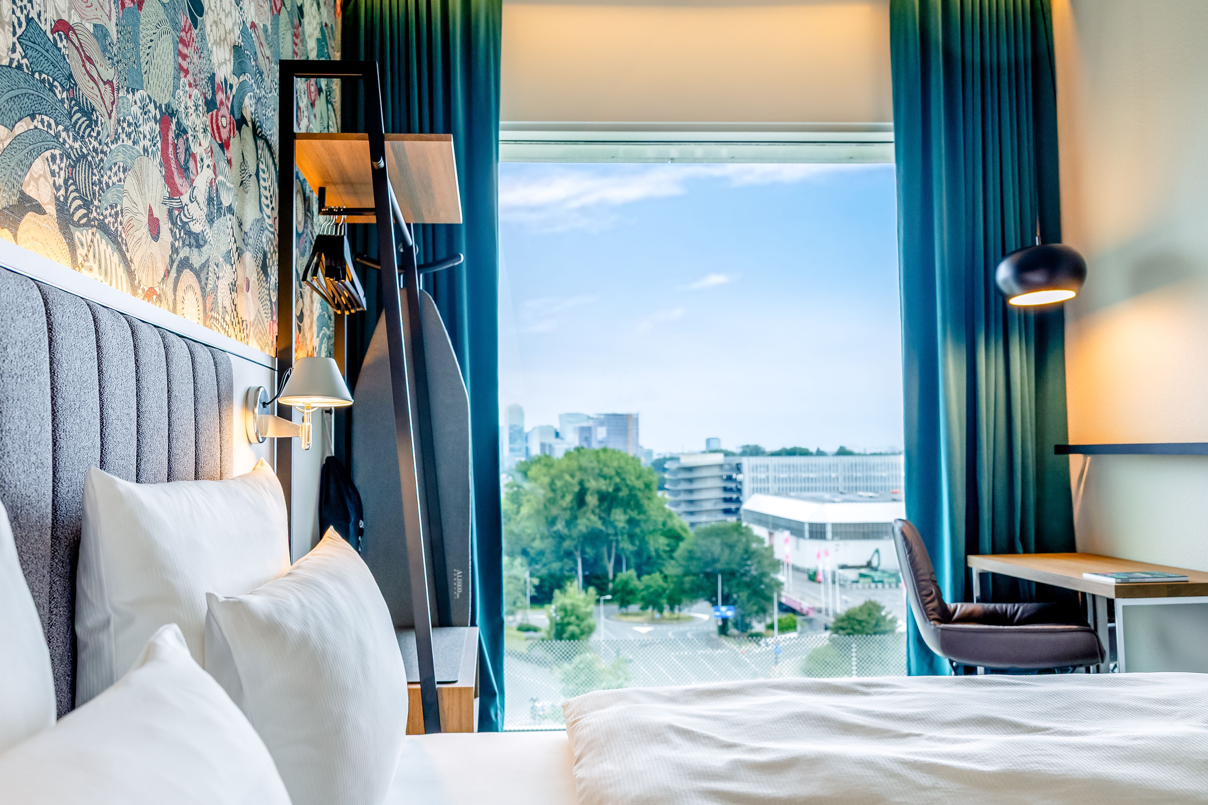 Helles Hotelzimmer mit großem Fenster, durch das man Stadtgebäude und Bäume sieht. Gemütliches Bett, Schreibtisch mit Stuhl und Vorhänge schaffen eine einladende Atmosphäre.