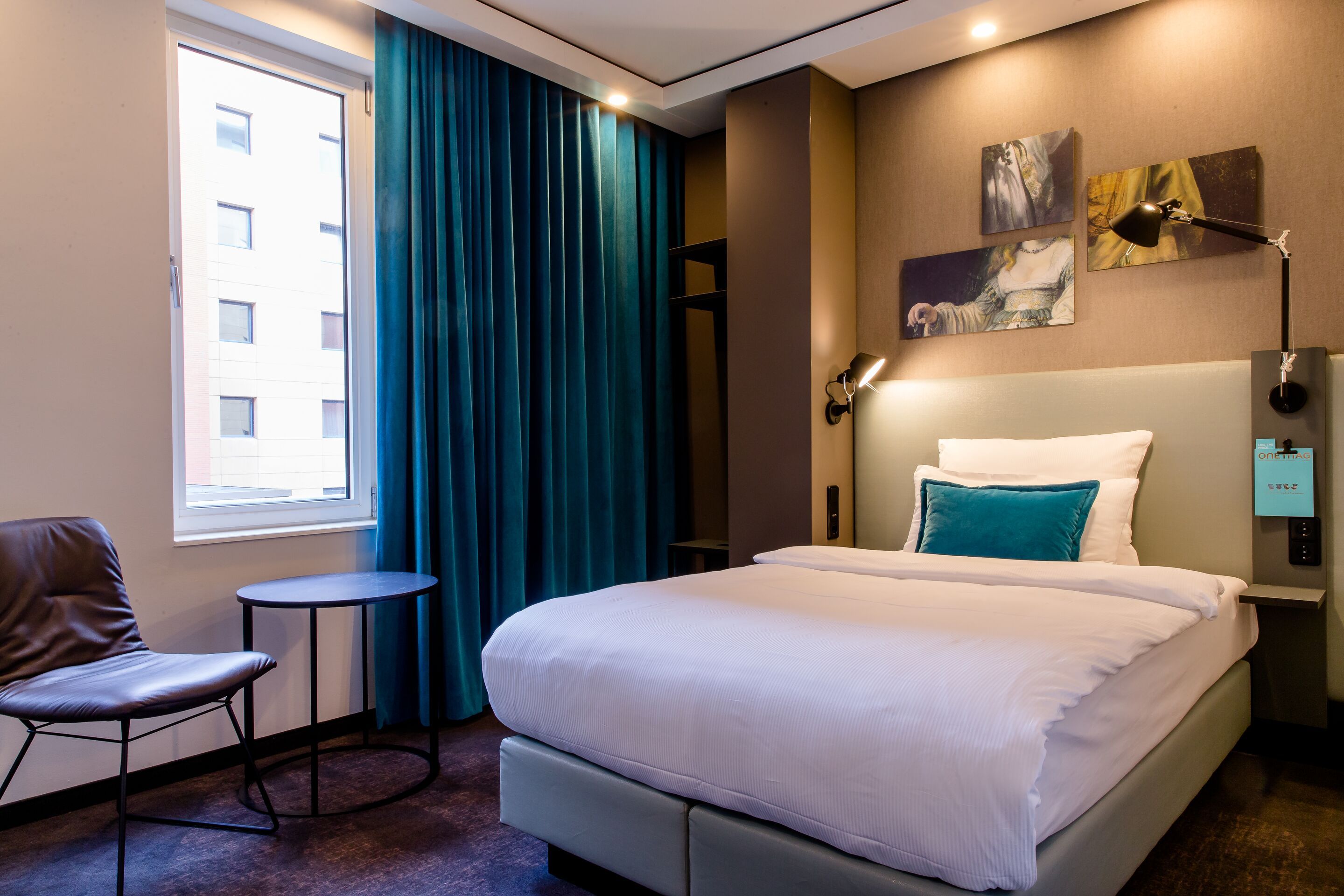 Modern eingerichtetes Hotelzimmer mit großem, bequemem Doppelbett, gemütlichem Sessel, Nachttischlampen und dunkelblauen Vorhängen vor einem Fenster, das Tageslicht hereinlässt. Die Atmosphäre wirkt ruhig und einladend.
