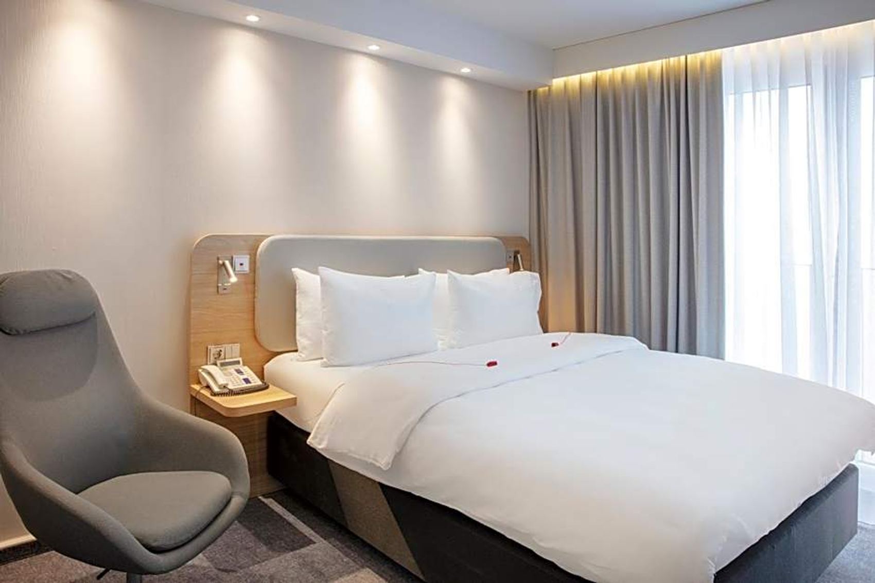 Modern eingerichtetes Hotelzimmer mit großem, weiß bezogenem Doppelbett, weicher Beleuchtung über dem Kopfteil, bequemen grauem Sessel und bodentiefen Fenstern mit hellen Vorhängen für viel Tageslicht.