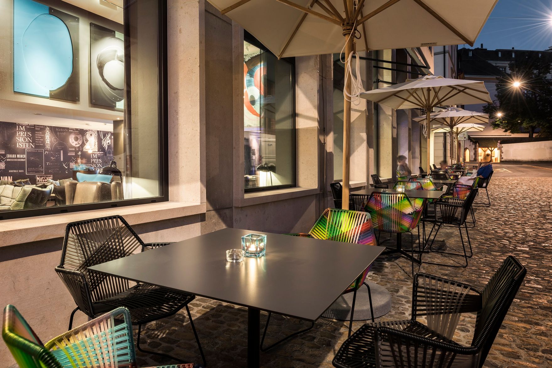 Hotel Basel Motel One Außenbereich mit Tischen und Stühlen sowie Sonnenschirmen bei Abend