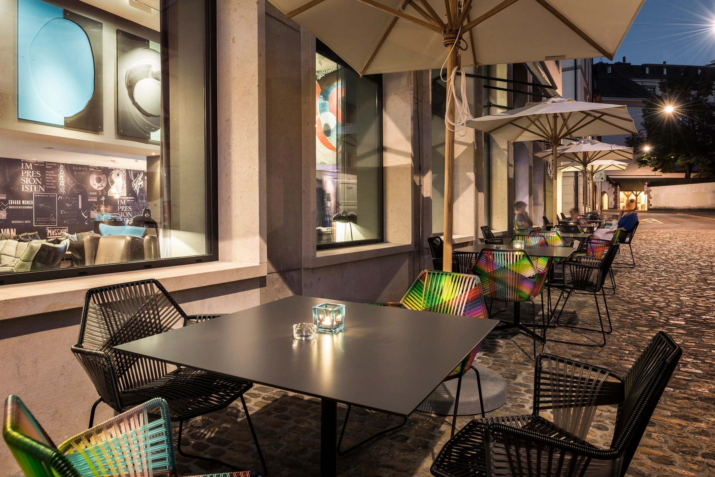 Hotel Basel Motel One Außenbereich mit Tischen und Stühlen sowie Sonnenschirmen bei Abend