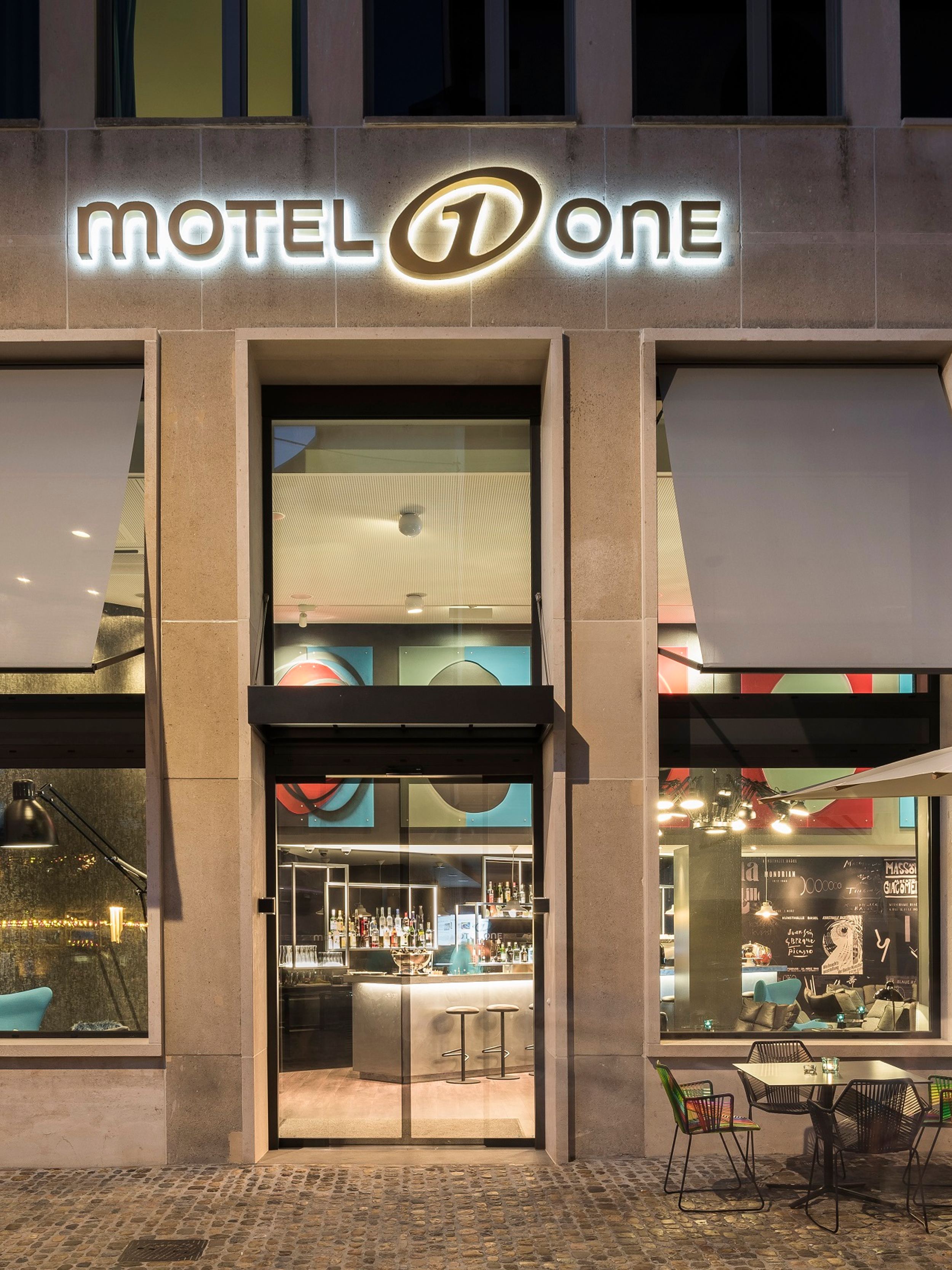 Hotel Basel Motel One Blick von außen auf den Eingangsbereich des Hotels mit Sitzgelegenheiten