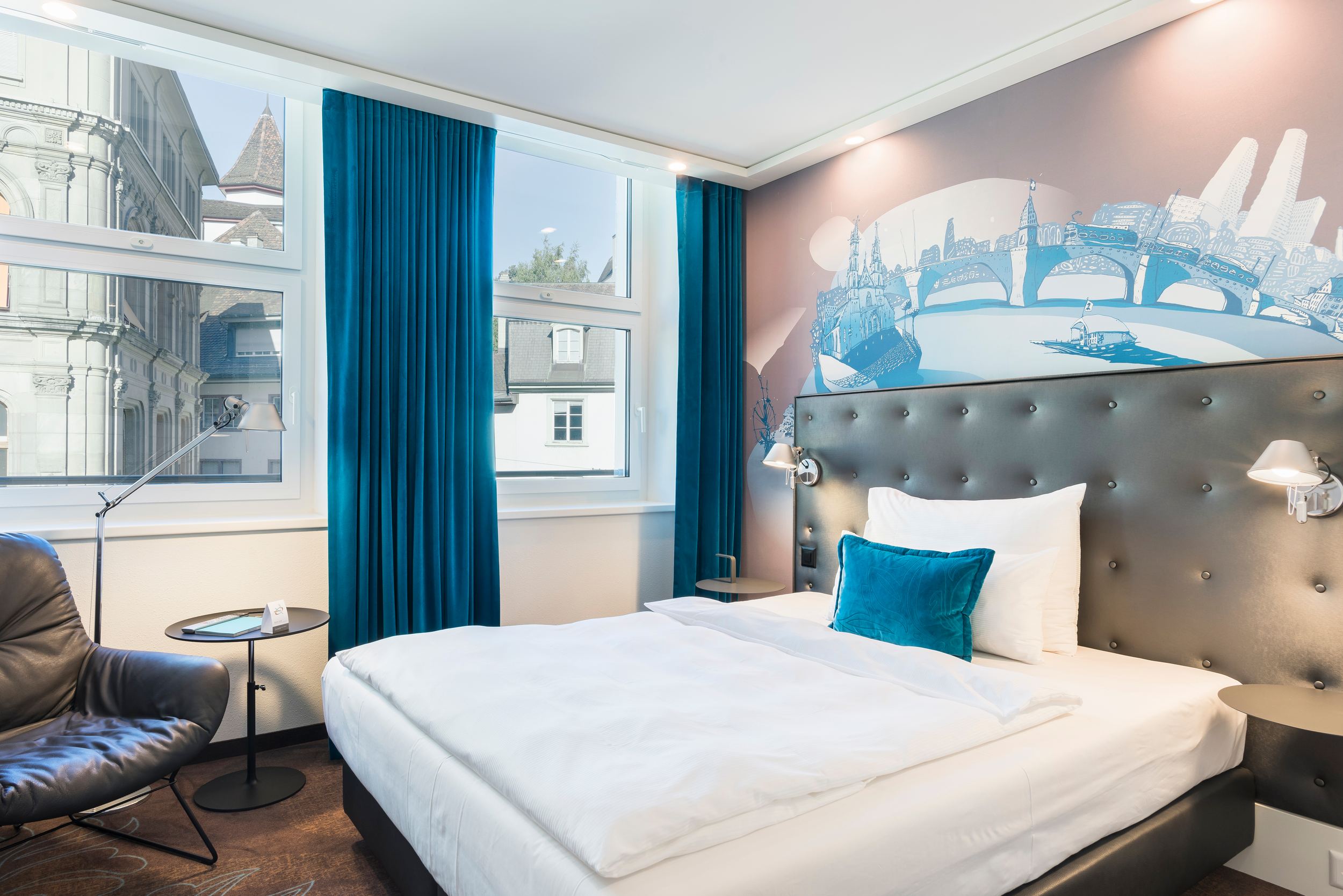 Designhotel Basel Motel One seitlicher Blick in ein helles Zimmer mit Bett und Sessel