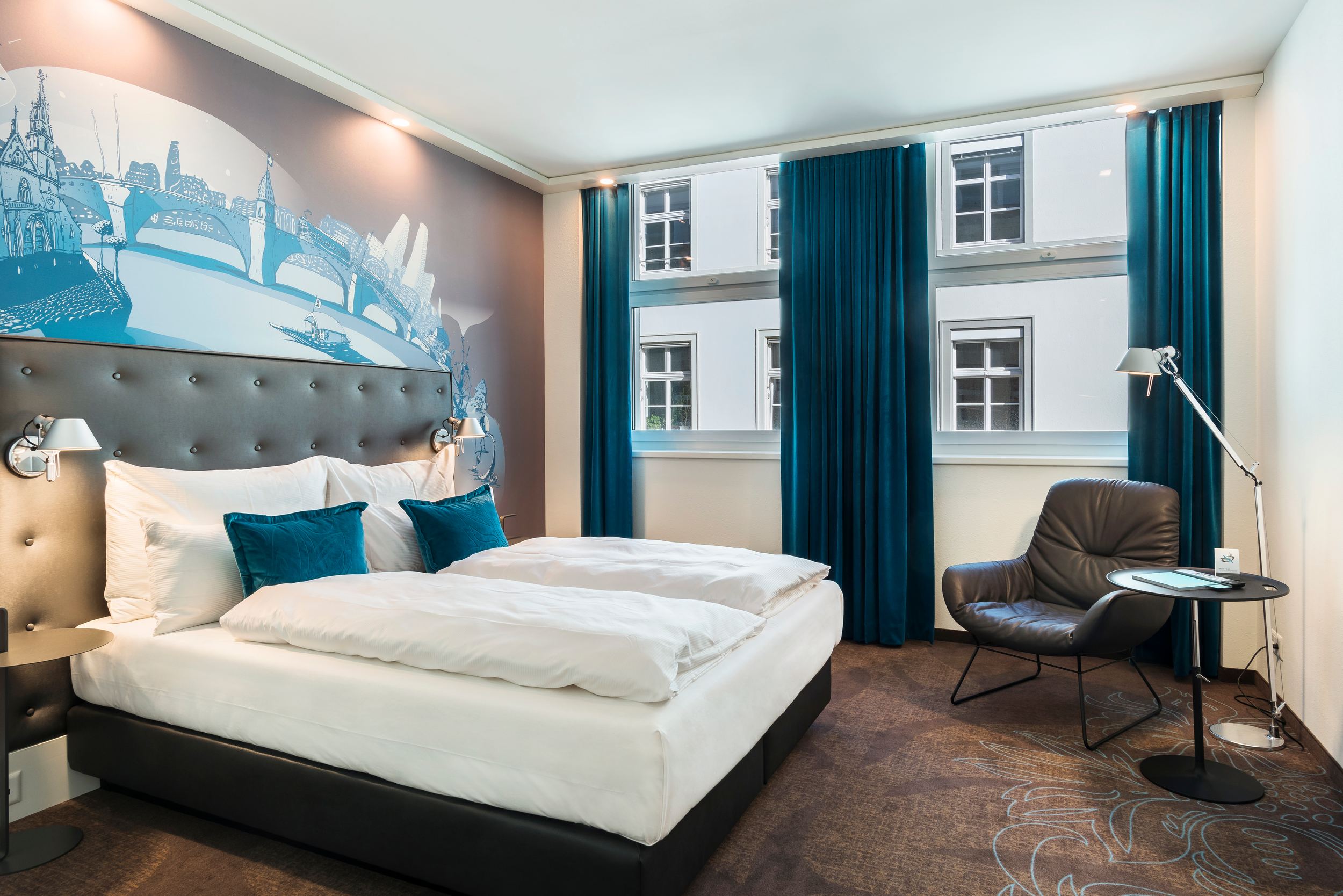 Designhotel Basel Motel One seitlicher Blick in ein helles Zimmer mit Doppelbett, Sessel, rundem Beistelltisch und Stehlampe