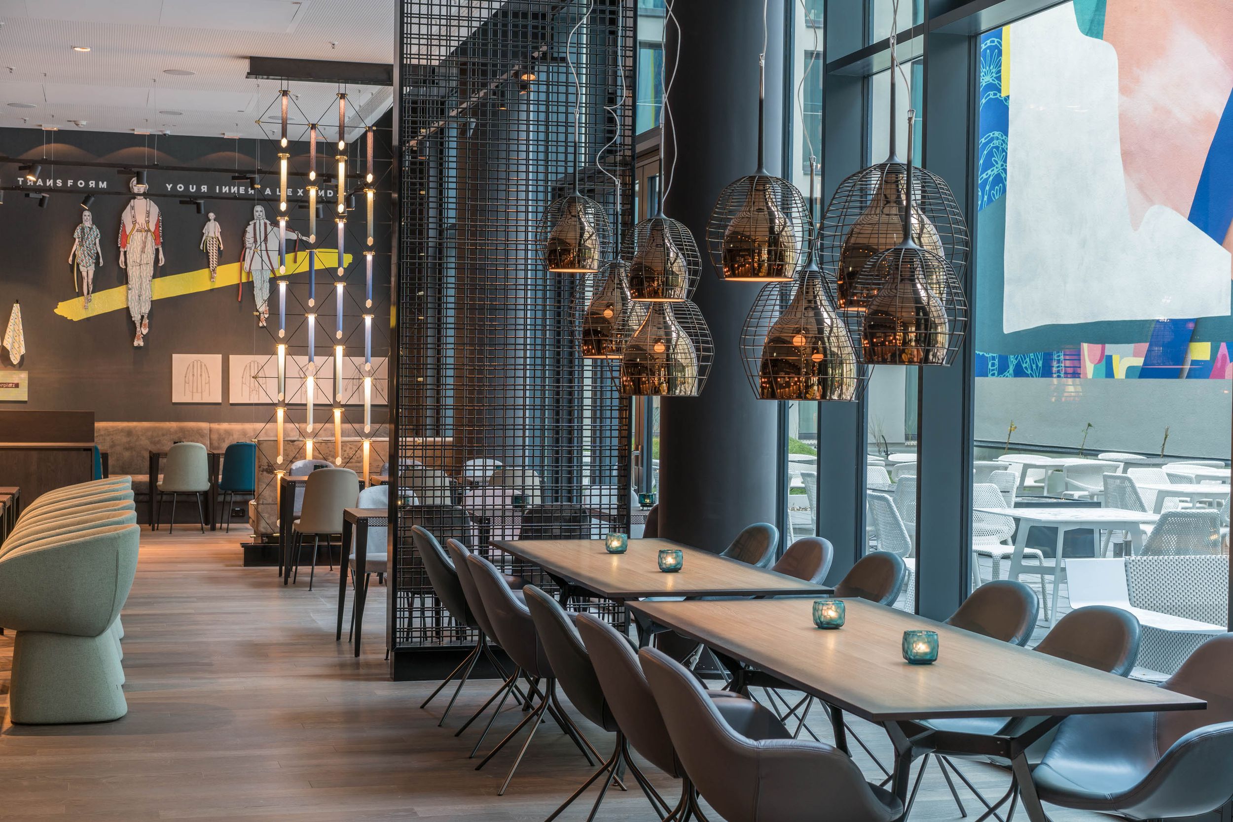 Hotel Berlin Alexanderplatz Motel One helle Lounge mit Tischen und Stühlen sowie große Fensterfront mit Blick auf den Außenbereich des Hotels mit weiteren Sitzgelegenheiten