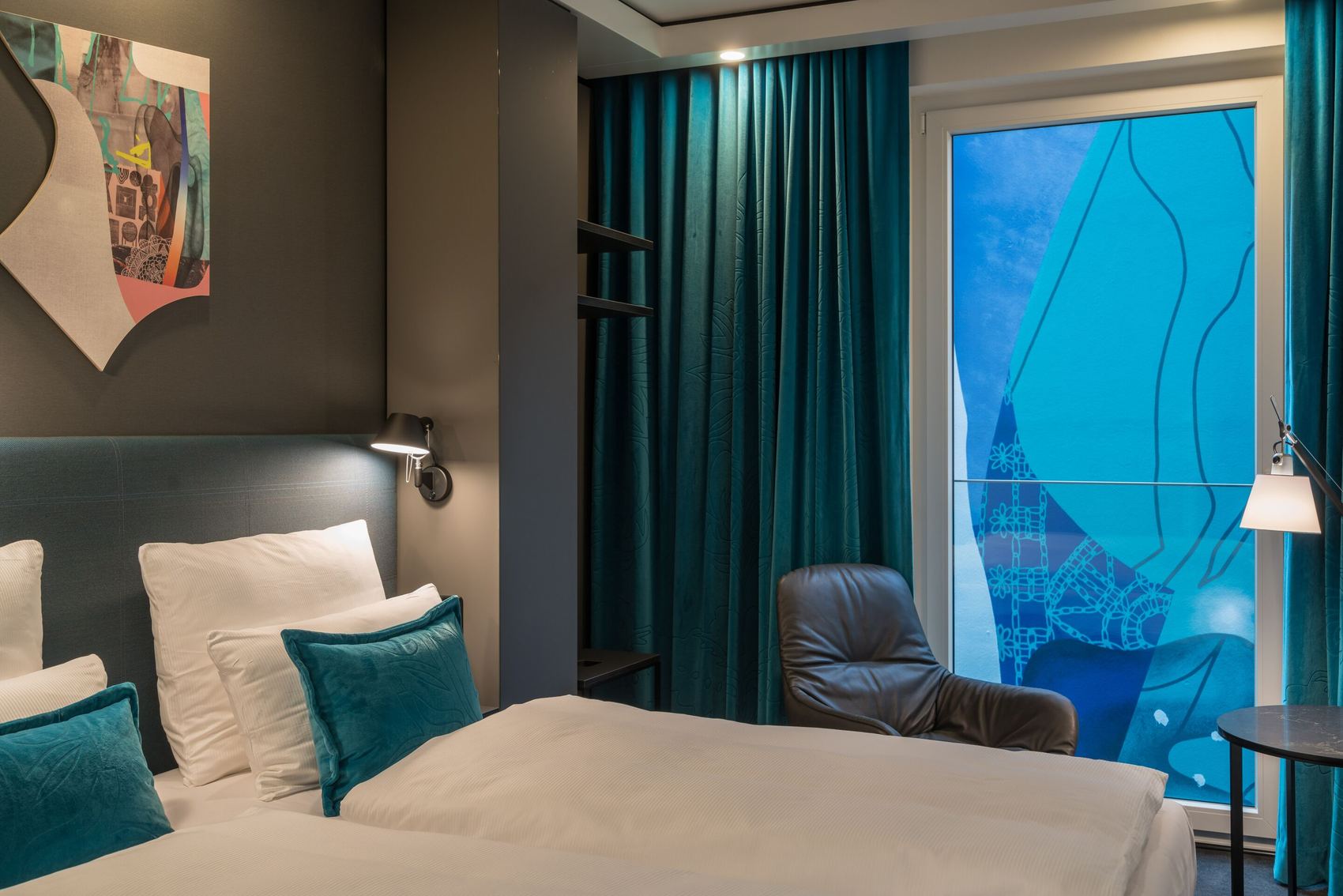 Modern eingerichtetes Hotelzimmer mit großem Doppelbett, dunklen Vorhängen, gemütlichem Sessel und einem großen Fenster mit dekorativer Glasgestaltung, das ruhige und komfortable Atmosphäre für erholsamen Aufenthalt bietet.