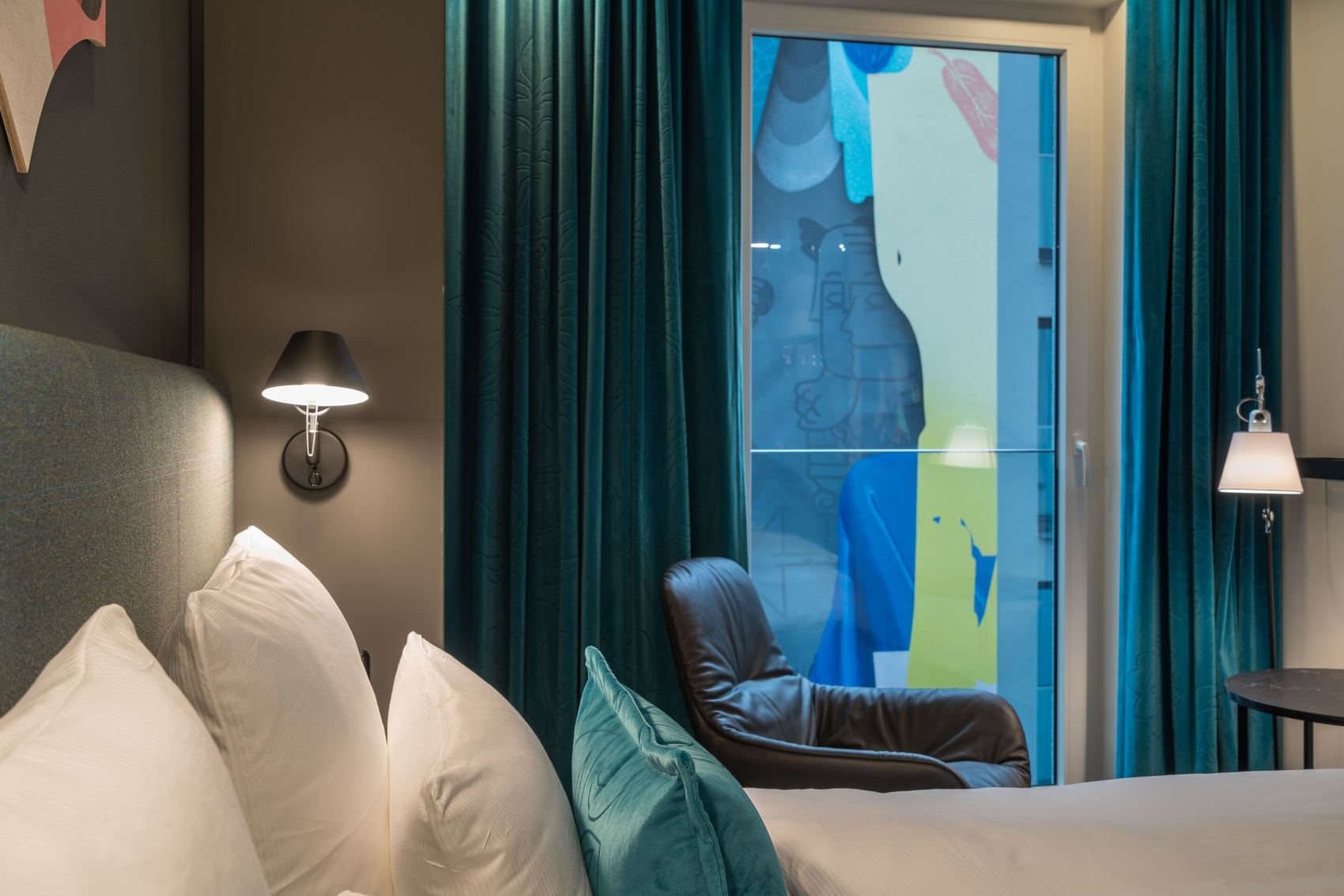 Modern eingerichtetes Hotelzimmer mit gemütlichem Bett, weichen Kissen, dunkelgrünen Vorhängen und Ledersessel vor einer großen Fensterfront mit Blick auf bunte, abstrakte Wandmalerei. Sanfte Beleuchtung sorgt für angenehme Atmosphäre.
