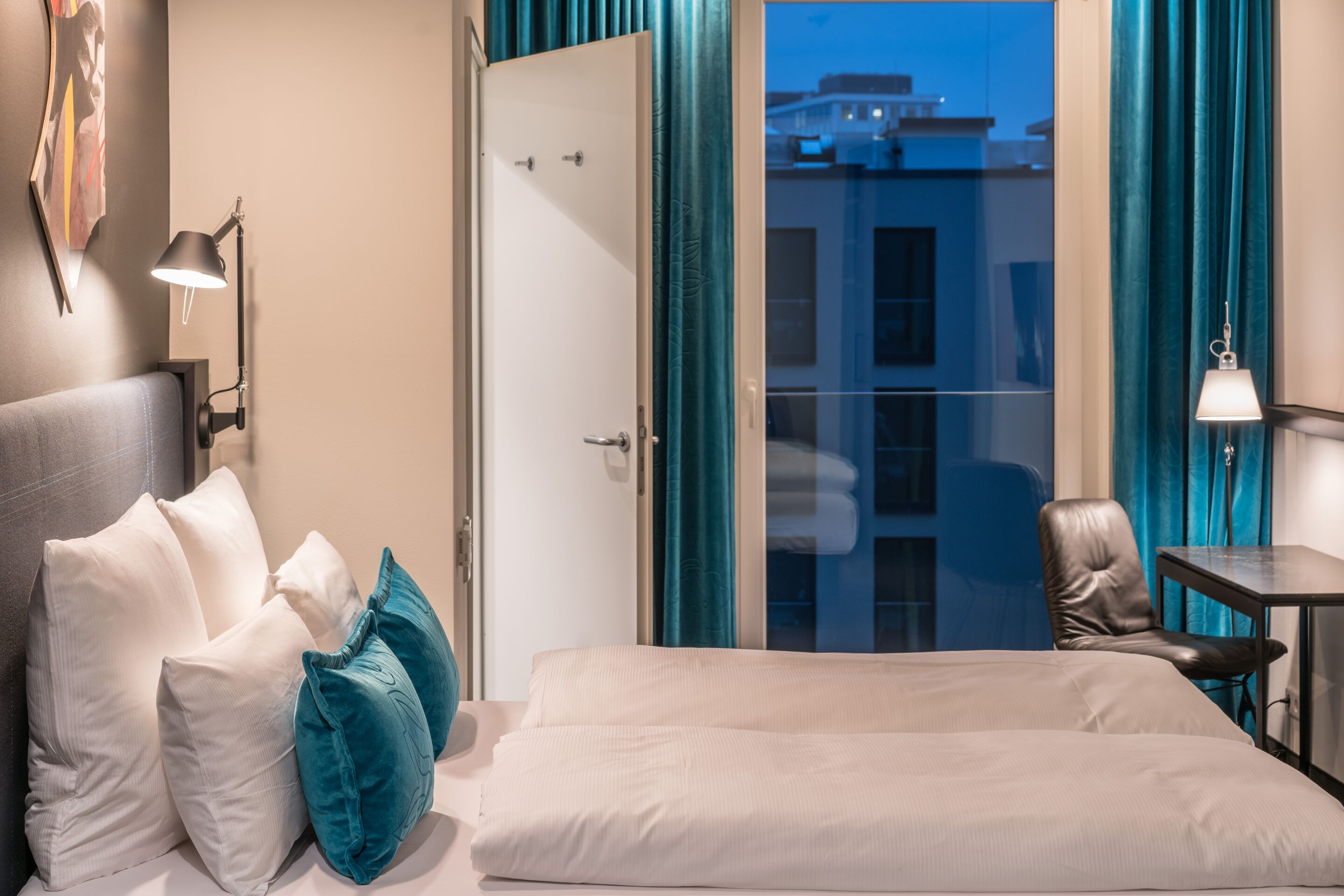Gemütliches Hotelzimmer mit großem Bett, weißen und türkisfarbenen Kissen, offenem Fenster mit Blick auf beleuchtete Gebäude und dunklen Vorhängen. Schreibtisch und Stuhl neben Fenster, warmes Licht schafft angenehme Atmosphäre.