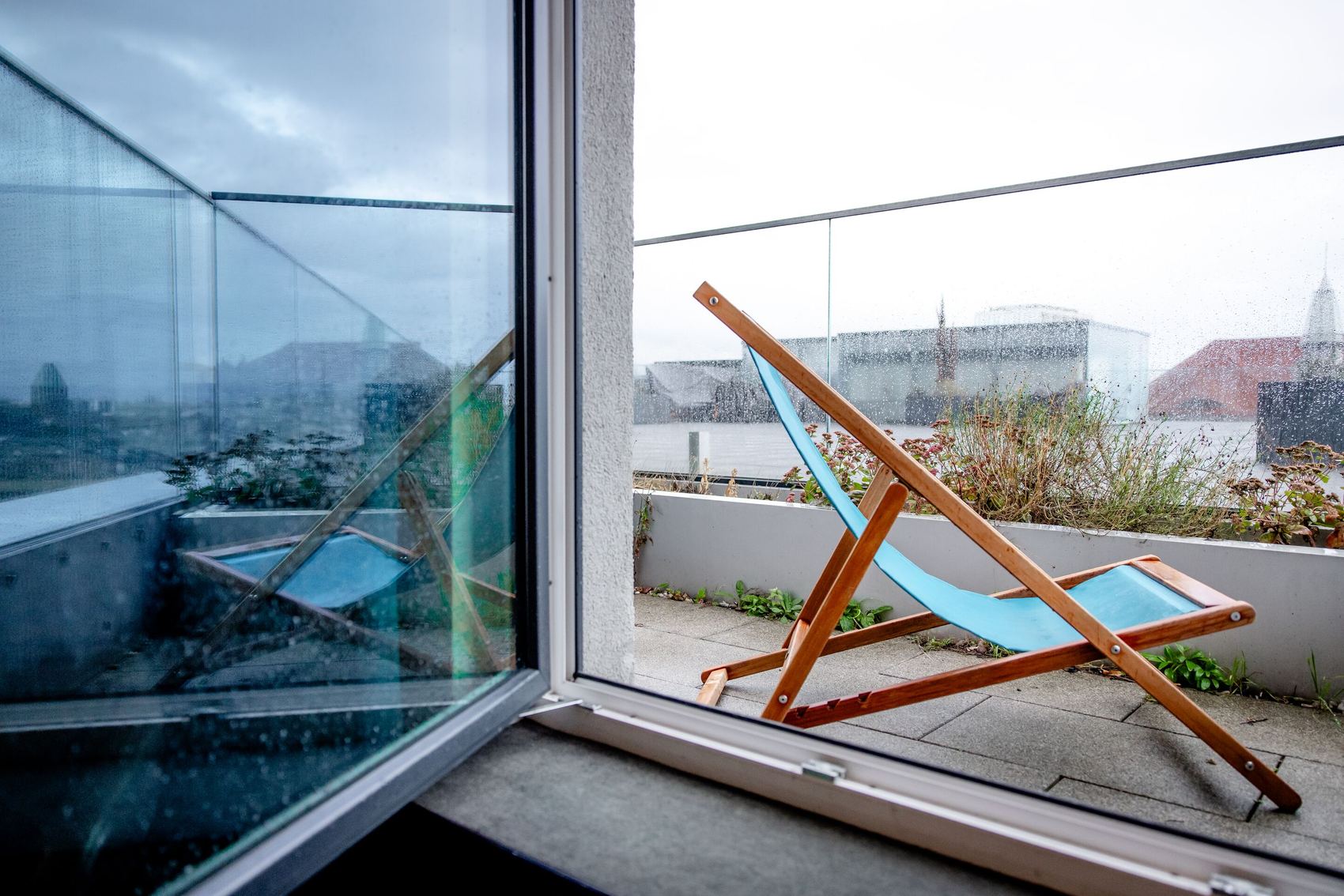 Blick von einem Zimmer auf einen Balkon mit einem blauen Liegestuhl, umgeben von Glasgeländern, bei bewölktem Himmel und Regen.