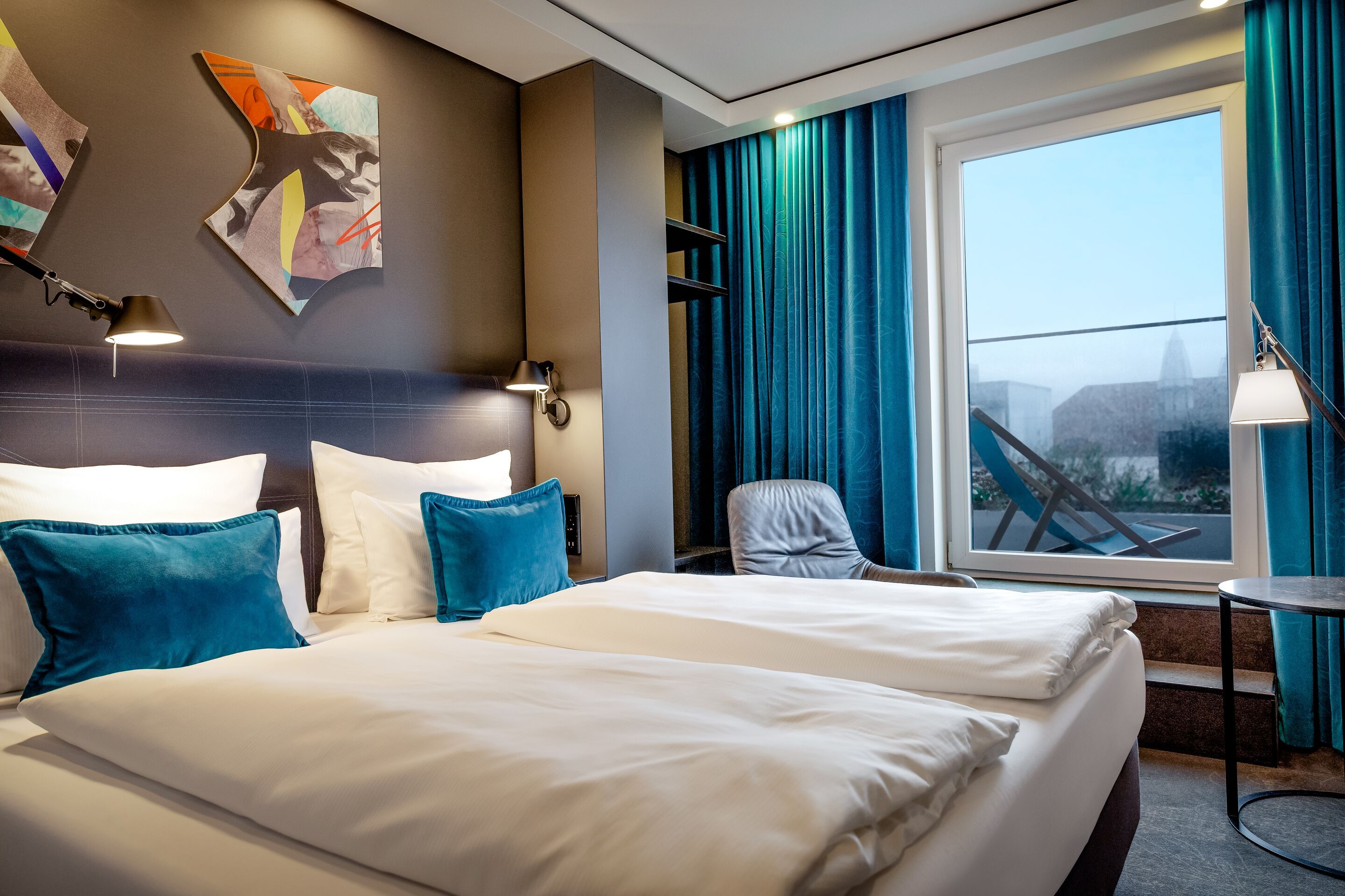 Modern eingerichtetes Hotelzimmer mit Queensize-Bett, blauen Akzentkissen, gemütlichem Lesesessel, großem Fenster mit Ausblick und stilvoller Beleuchtung für entspannte Aufenthalte.