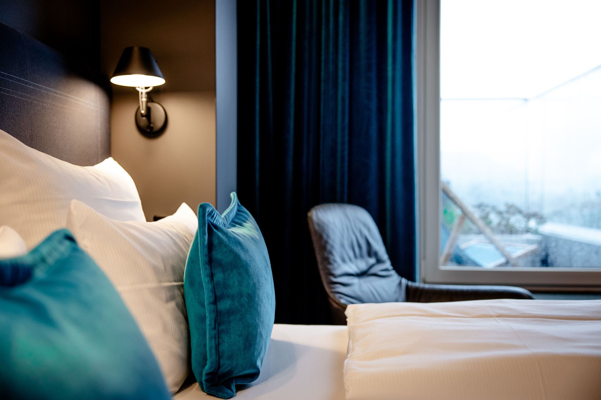 Gemütliches Hotelzimmer mit großem, komfortablem Bett, weichen blauen Kissen, gedämpftem Licht und einem bequemen Sessel am Fenster mit Ausblick. Ideal für erholsame Nächte und entspannte Momente.