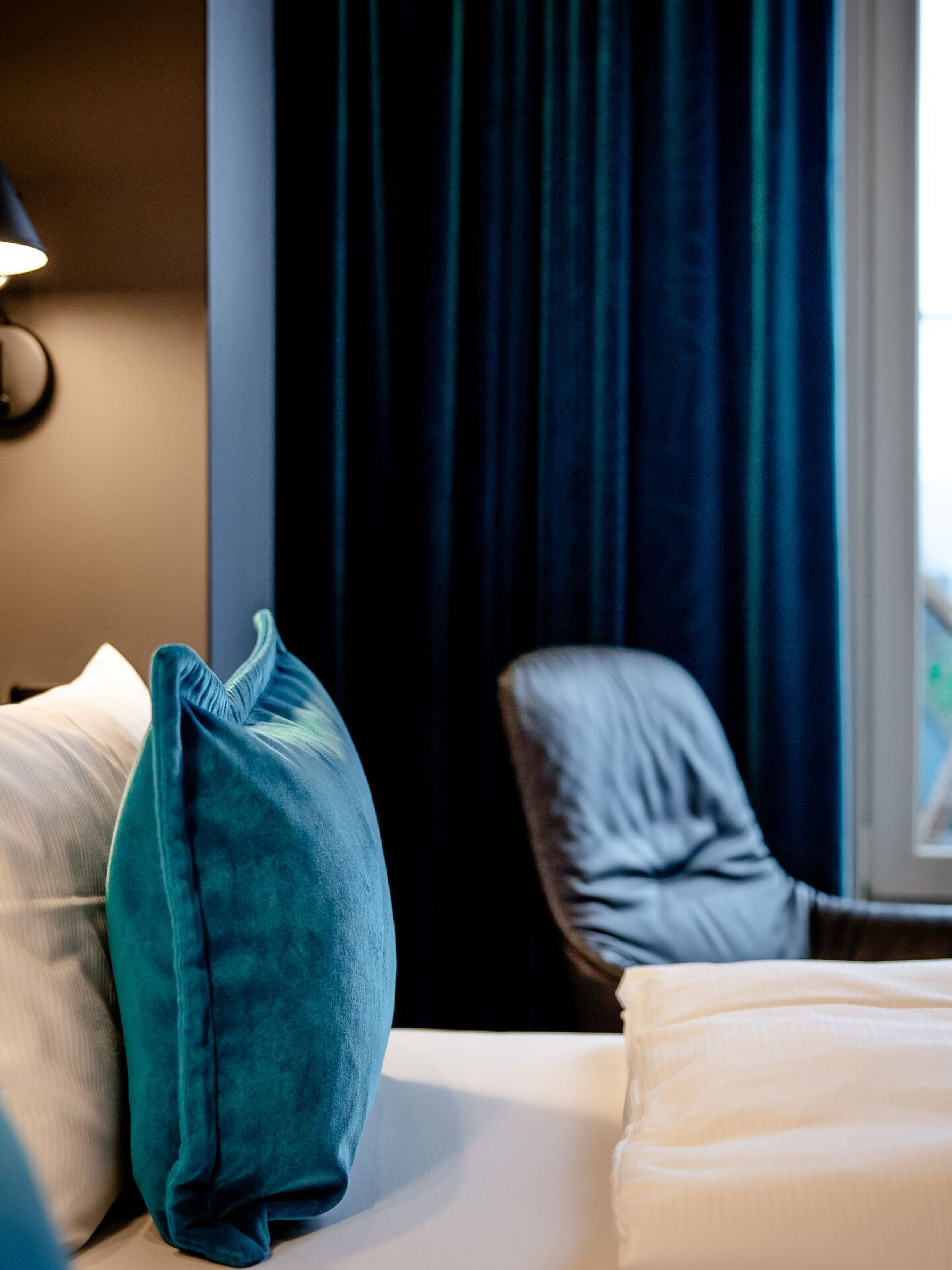 Gemütliches Hotelzimmer mit großem, komfortablem Bett, weichen blauen Kissen, gedämpftem Licht und einem bequemen Sessel am Fenster mit Ausblick. Ideal für erholsame Nächte und entspannte Momente.