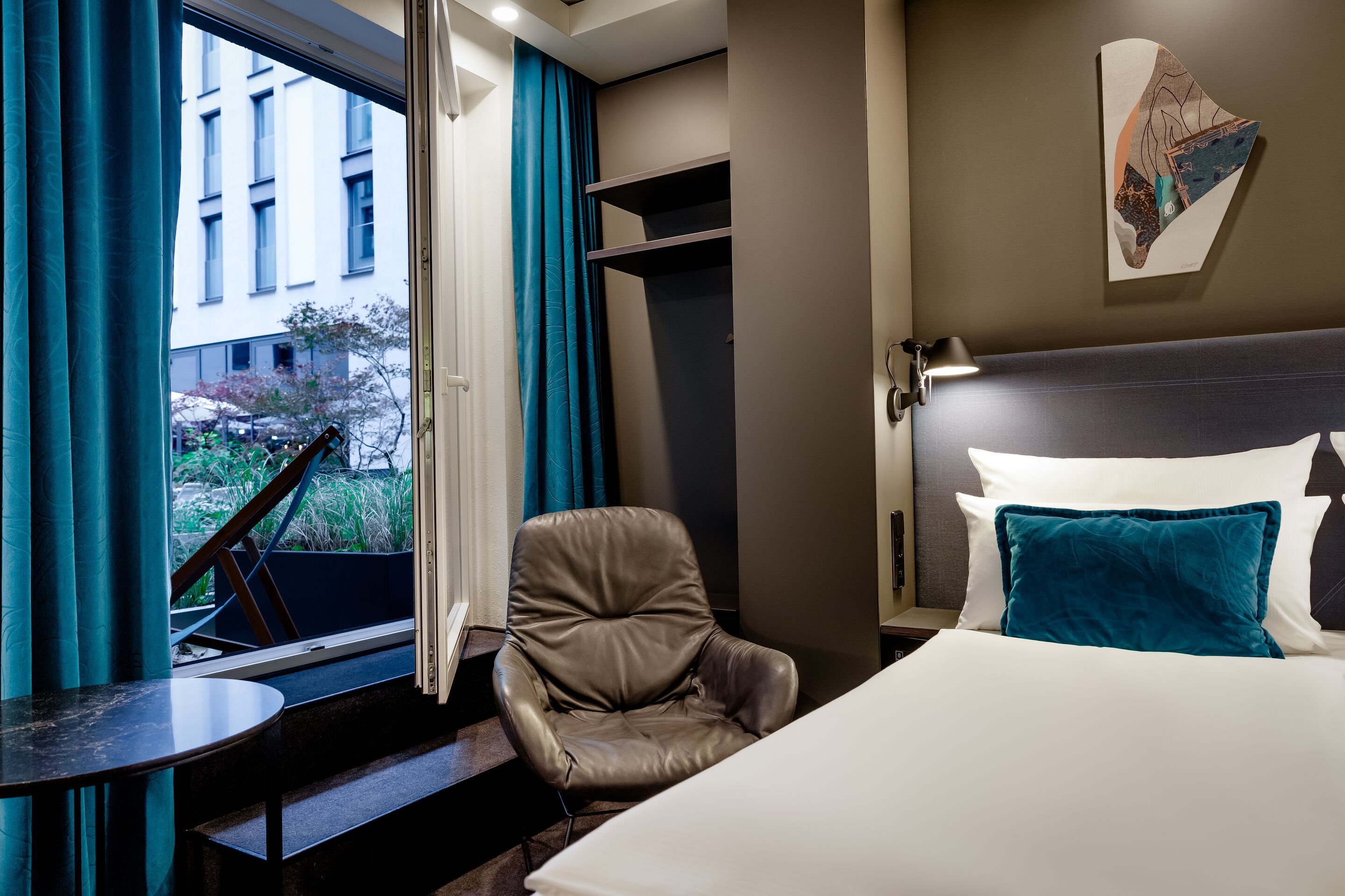Modern eingerichtetes Hotelzimmer mit großem Bett, bequemen Sessel und Zugang zu einem Balkon mit Blick auf eine ruhige Innenhofanlage. Helle Farben und stilvolle Dekoration schaffen eine entspannte Atmosphäre.