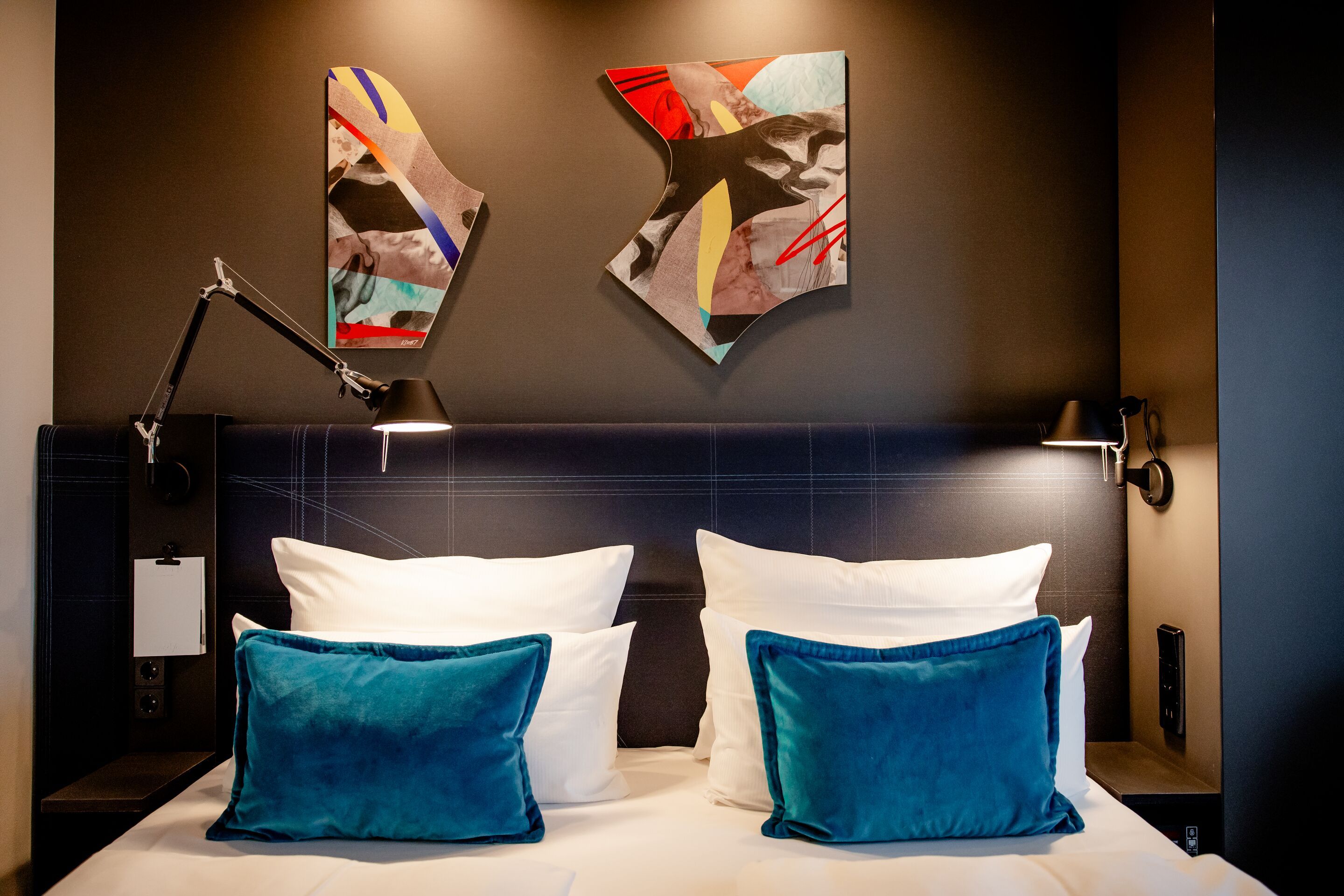 Modern eingerichtetes Doppelzimmer mit gemütlichem Bett, weißen Kissen und zwei türkisblauen Zierkissen, stimmungsvolle Wandbeleuchtung und abstrakter Kunst an der Wand für ein entspanntes Ambiente.