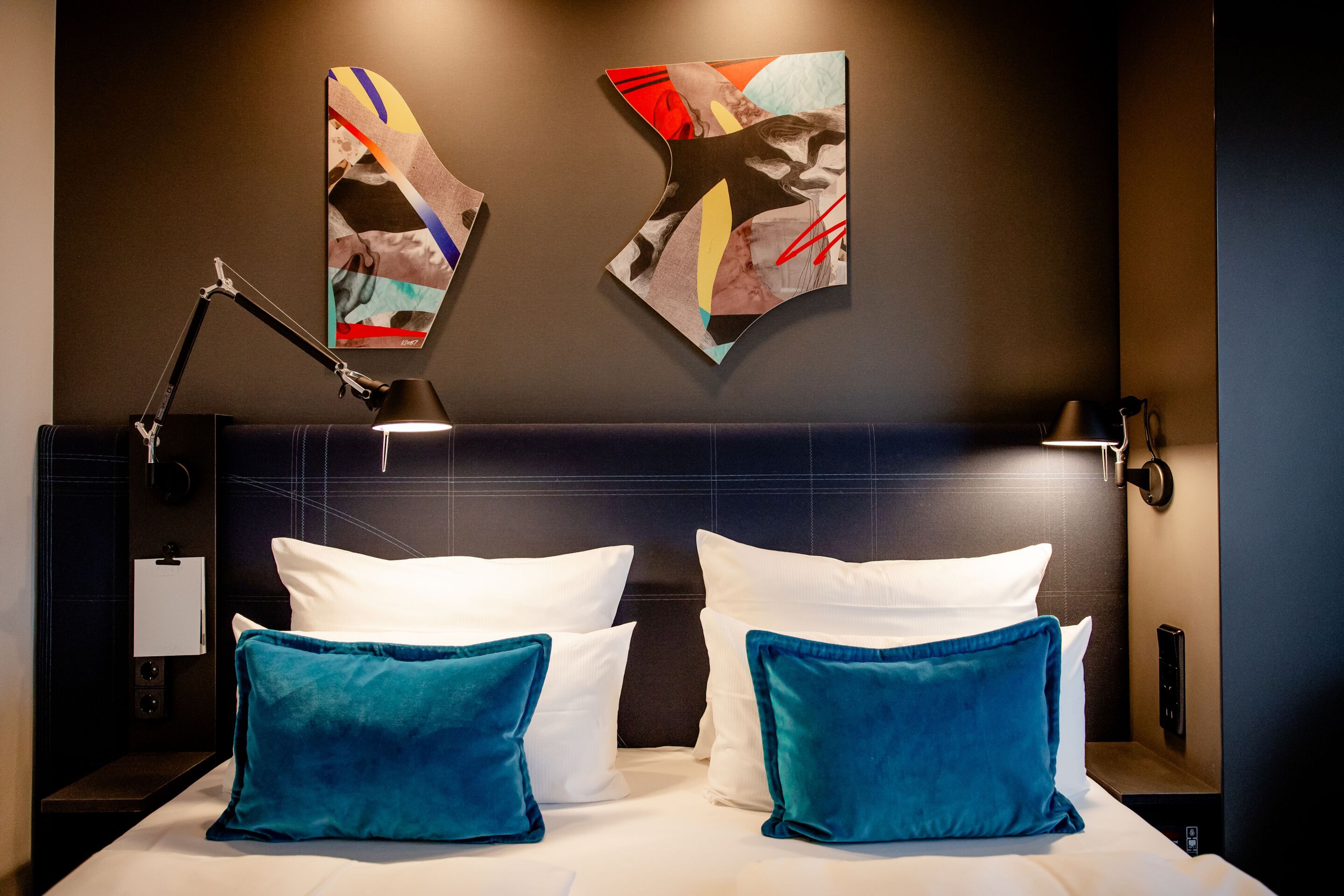 Modern eingerichtetes Doppelzimmer mit gemütlichem Bett, weißen Kissen und zwei türkisblauen Zierkissen, stimmungsvolle Wandbeleuchtung und abstrakter Kunst an der Wand für ein entspanntes Ambiente. Modern eingerichtetes Doppelzimmer mit gemütlichem Bett, weißen Kissen und zwei türkisblauen Zierkissen, stimmungsvolle Wandbeleuchtung und abstrakter Kunst an der Wand für ein entspanntes Ambiente.