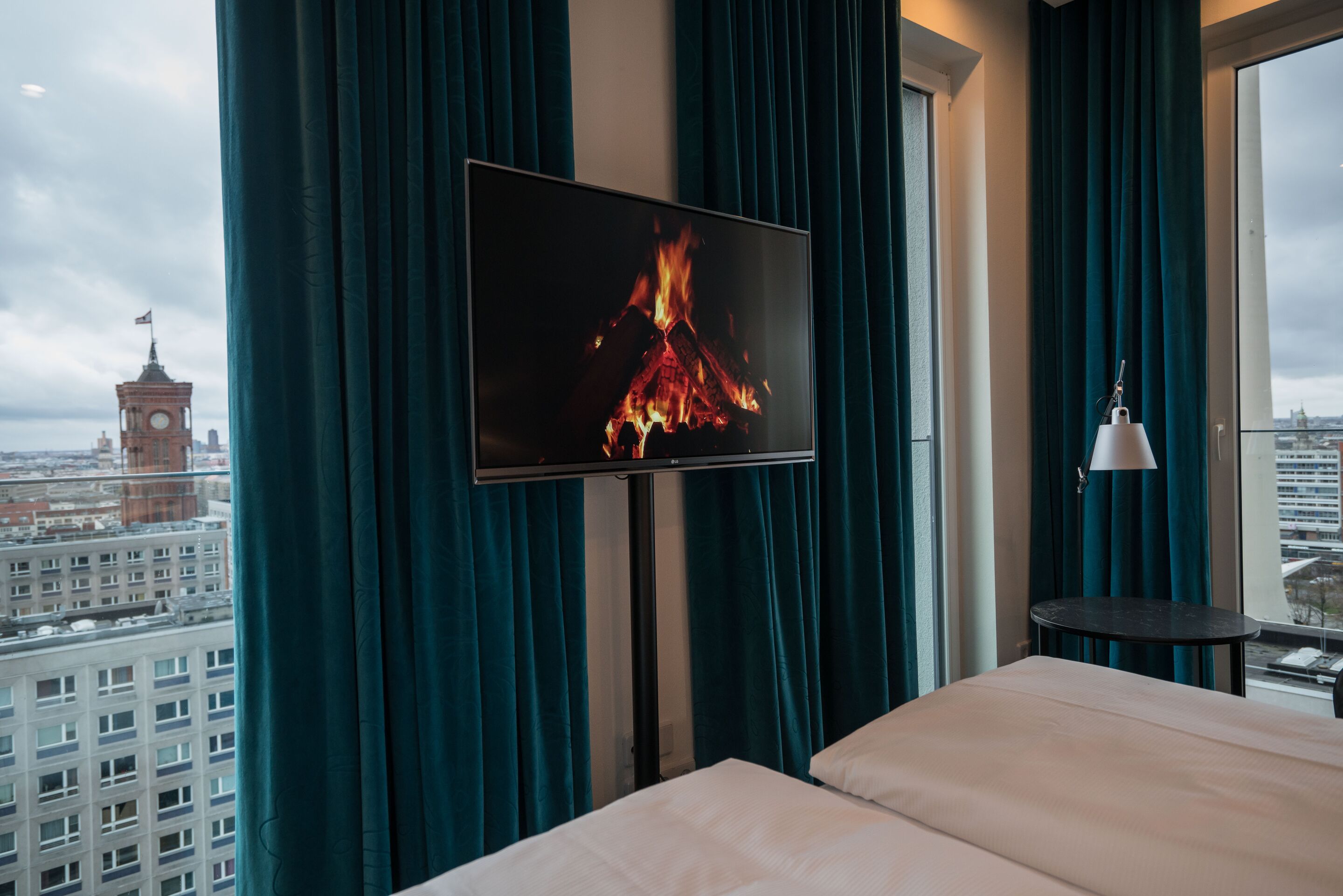 Blick aus einem Hotelzimmer mit gemütlichem Doppelbett, Fernseher zeigt flackerndes Kaminfeuer, große Fenster mit Blick auf Stadt und historischen Turm, dunkelgrüne Vorhänge schaffen behagliche Atmosphäre.