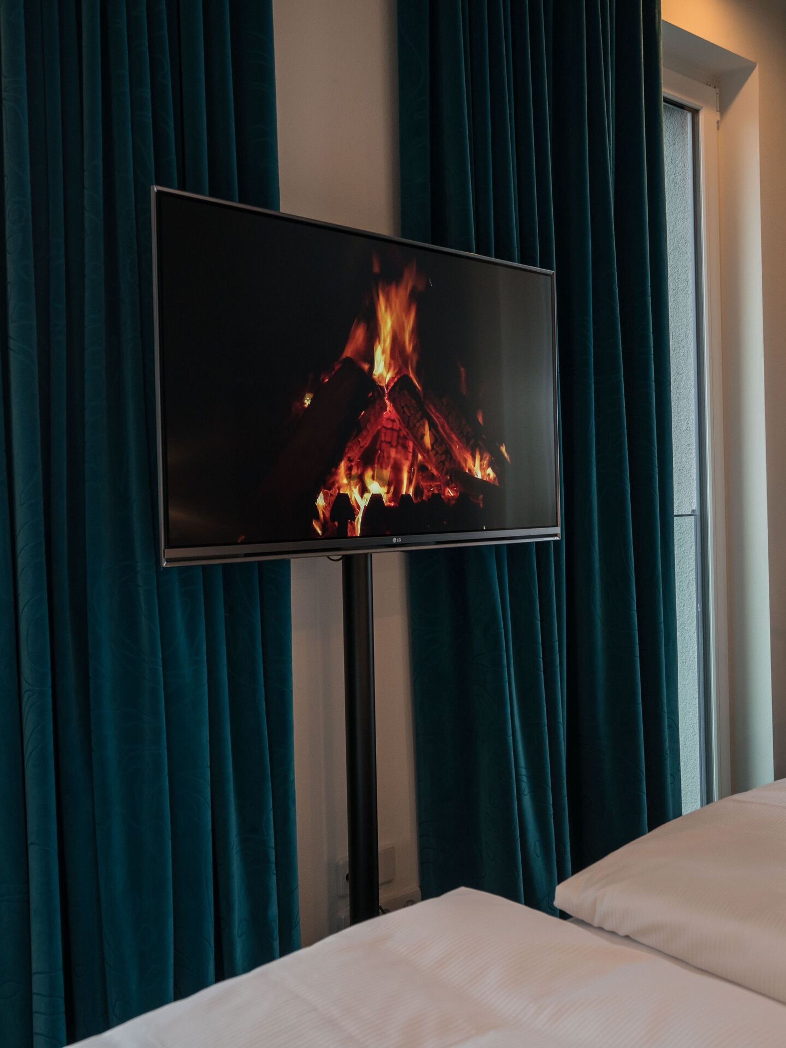 Blick aus einem Hotelzimmer mit gemütlichem Doppelbett, Fernseher zeigt flackerndes Kaminfeuer, große Fenster mit Blick auf Stadt und historischen Turm, dunkelgrüne Vorhänge schaffen behagliche Atmosphäre.