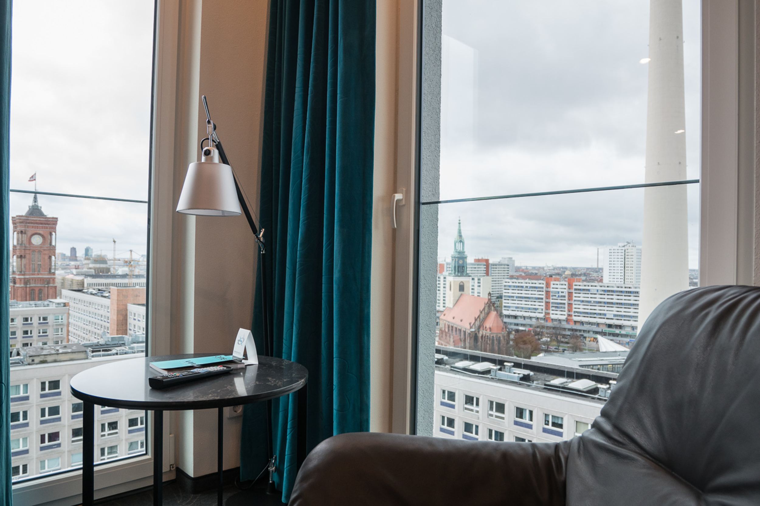 Hotel Berlin Alexanderplatz Motel One Eckzimmer mit zwei bodentiefen Fenstern und einem Sessel sowie Blick auf die Dächer der Stadt und den Fernsehturm