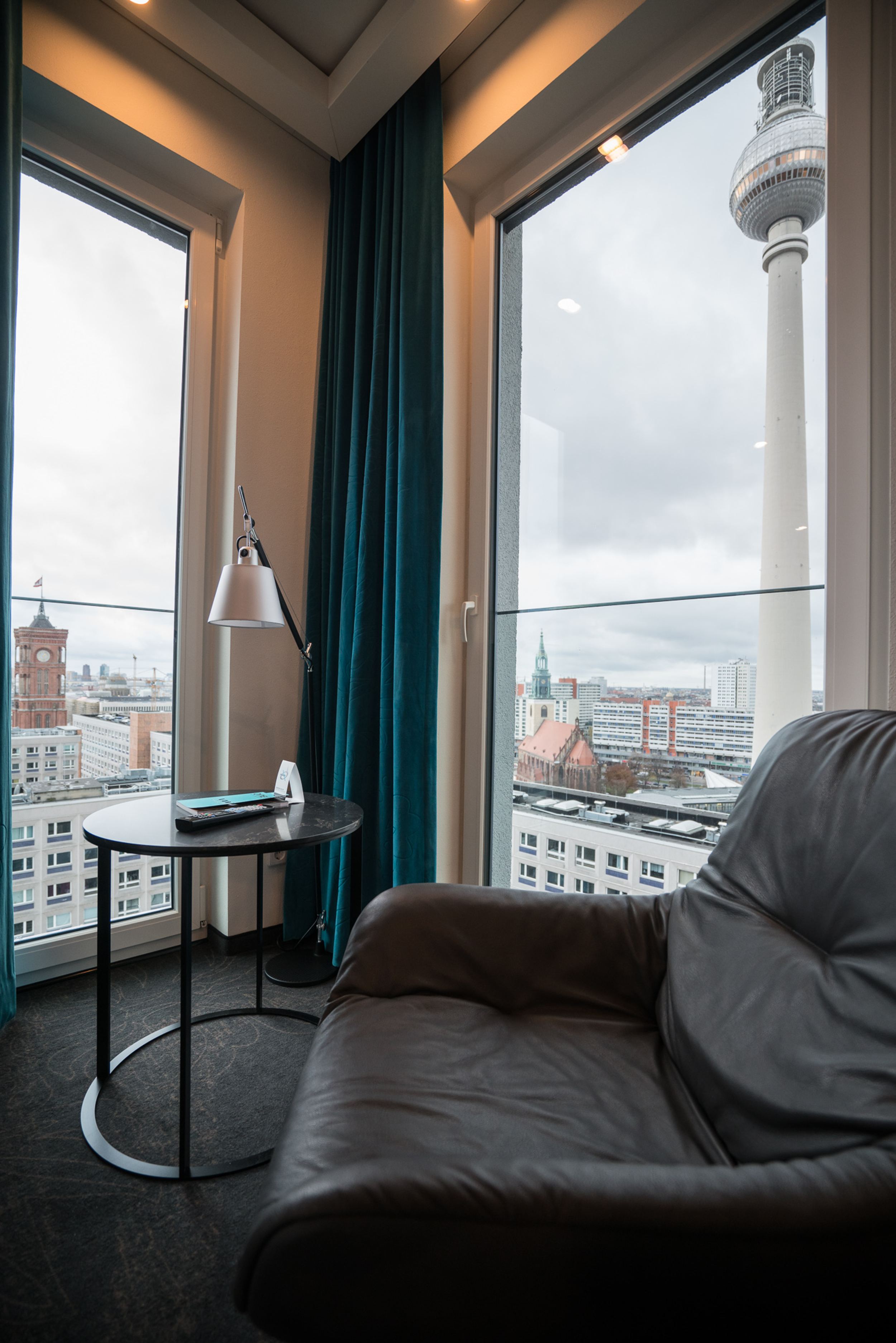 Hotel Berlin Alexanderplatz Motel One Eckzimmer mit zwei bodentiefen Fenstern und einem Sessel sowie Blick auf die Dächer der Stadt und den Fernsehturm