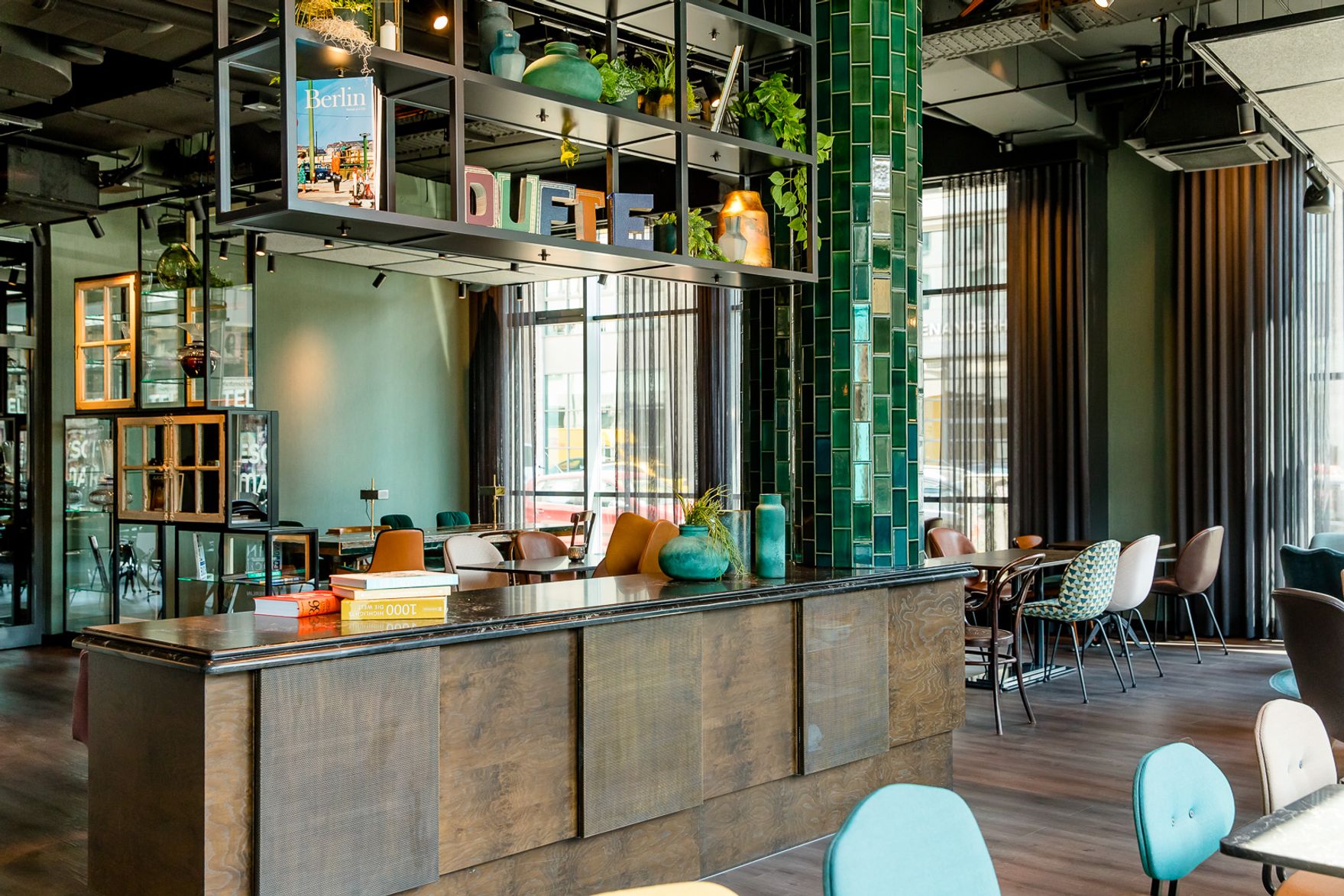 Hotel Berlin Hackescher Markt Motel One lichtdurchflutete Lounge mit mehreren Tischen und Stühlen sowie ein offenes Regal mit Dekoration in der Mitte des Raumes