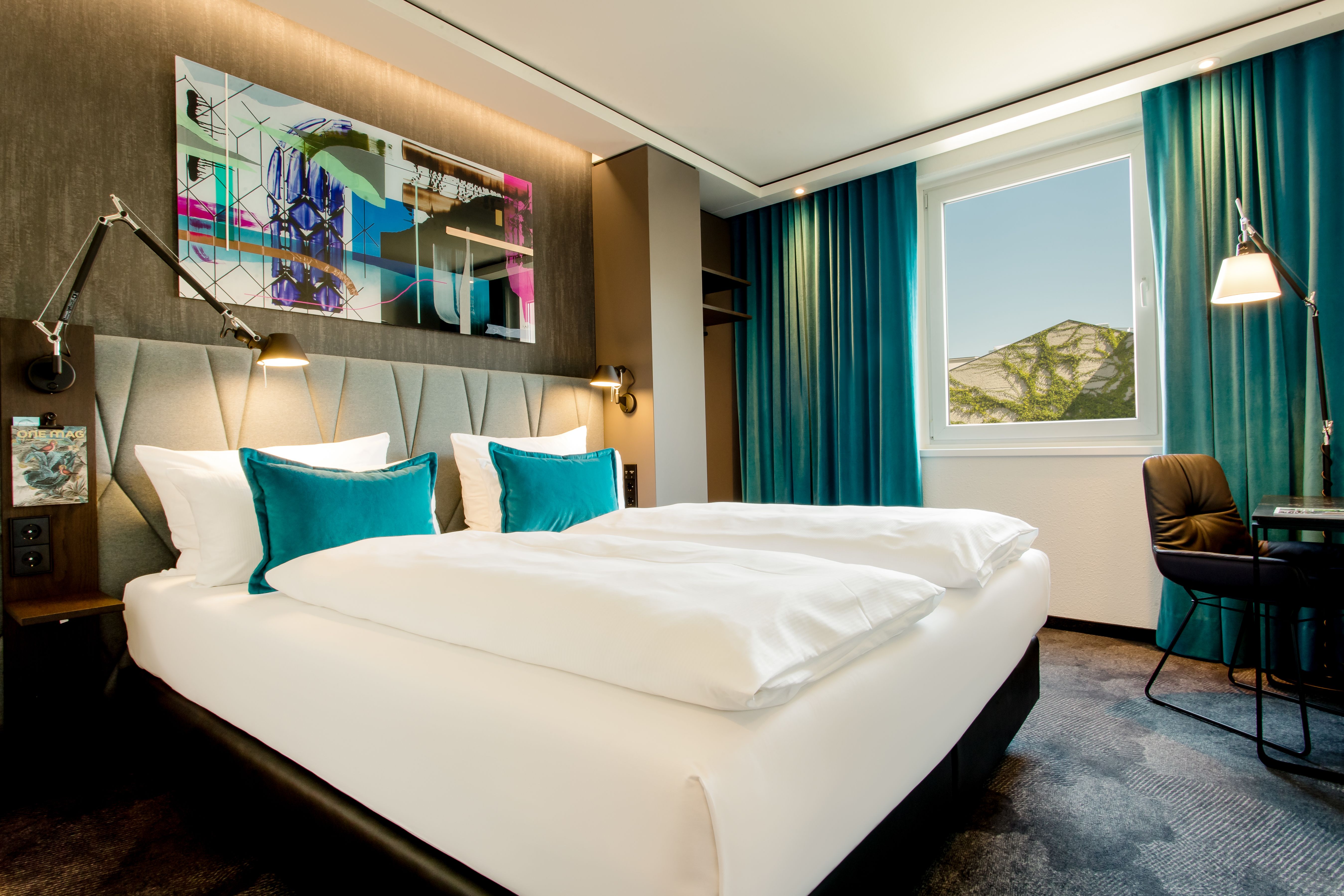 Hotel Berlin Hackescher Markt Motel One helles Zimmer mit komfortablem Doppelbett sowie Tisch, Stuhl und Stehlampe in der Ecke des Raumes