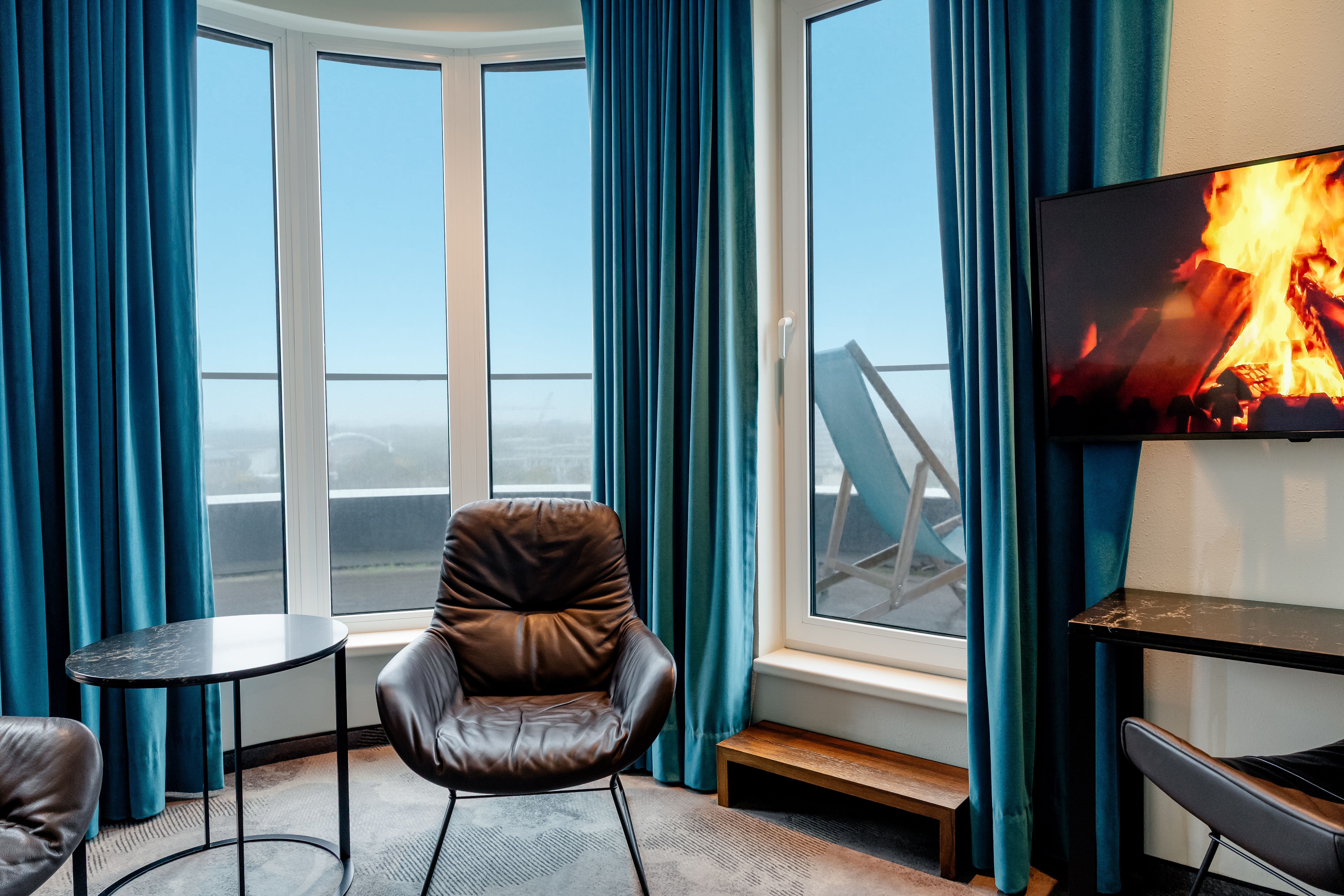 Hotel Motel One Berlin-Hauptbahnhof Gemütlicher Sitzbereich mit Ledersessel vor großen Fenstern, die auf eine Terrasse mit Liegestuhl und weitem Blick ins Freie führen. Im Raum sorgt ein Bildschirm mit Kaminfeuer für warme Atmosphäre.