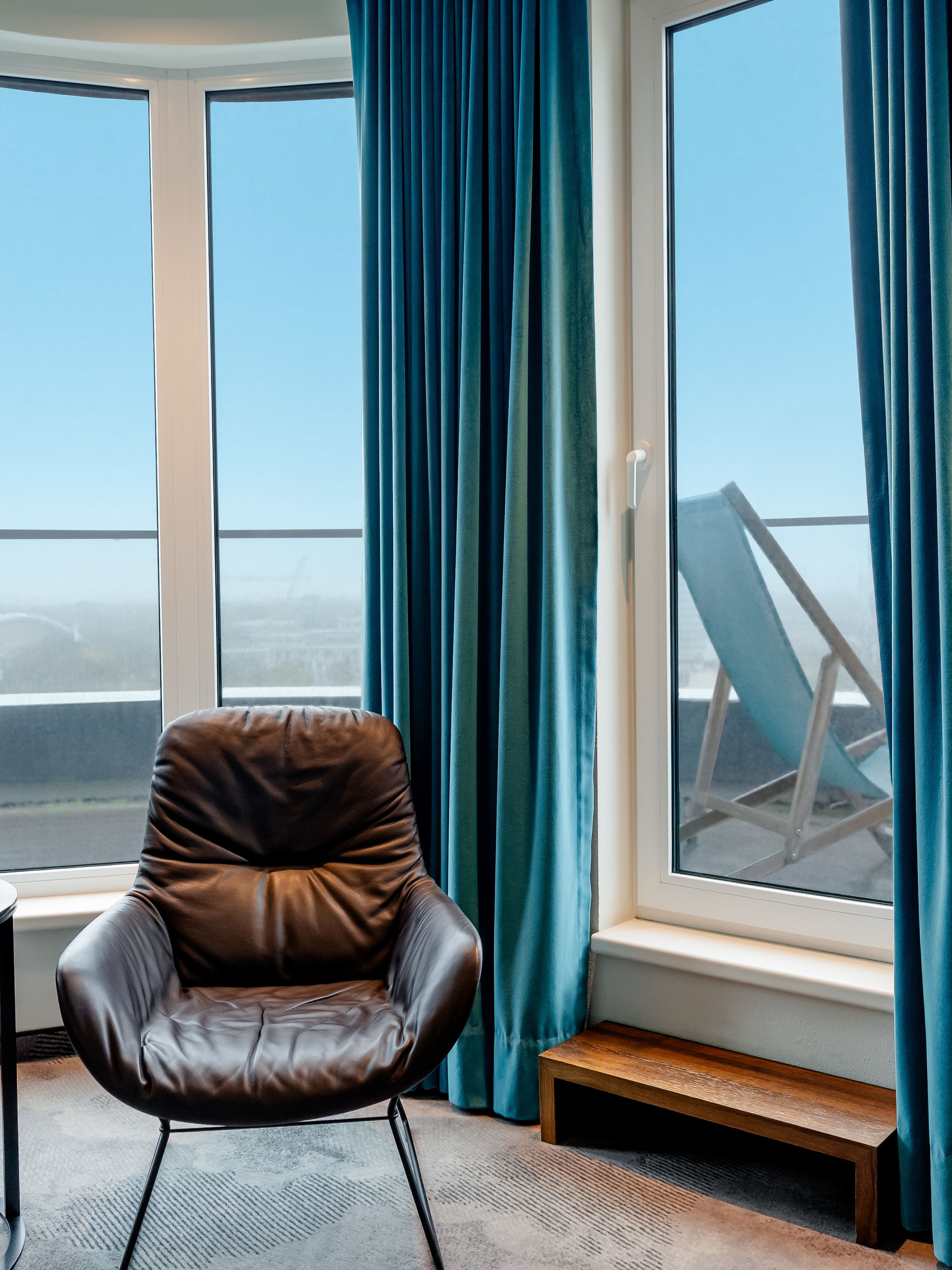 Hotel Motel One Berlin-Hauptbahnhof Gemütlicher Sitzbereich mit Ledersessel vor großen Fenstern, die auf eine Terrasse mit Liegestuhl und weitem Blick ins Freie führen. Im Raum sorgt ein Bildschirm mit Kaminfeuer für warme Atmosphäre.