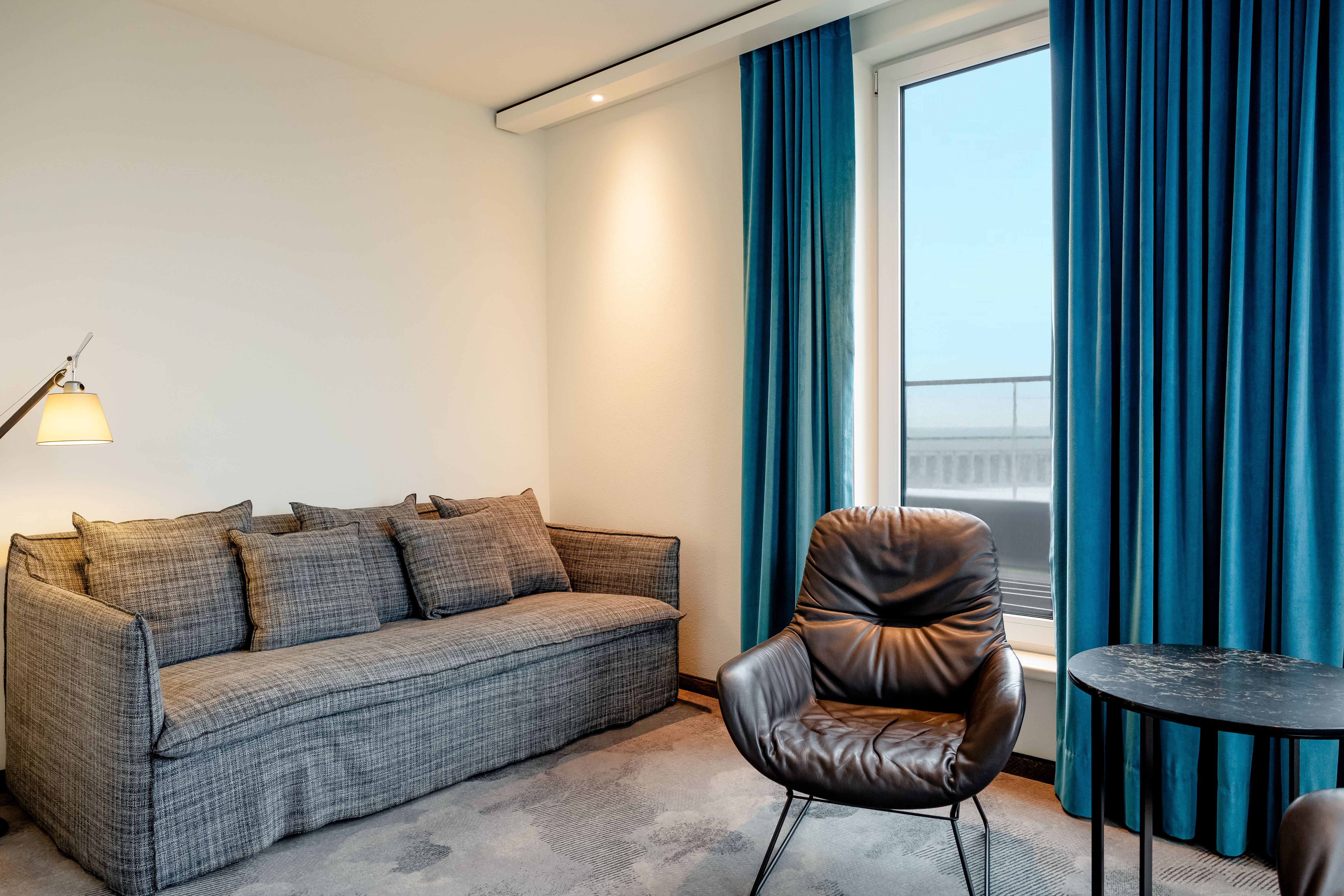Hotel Motel One Berlin-Hauptbahnhof Gemütliche Sitzecke mit grauem Sofa, schwarzem Leder-Sessel und großem Fenster mit blauen Vorhängen, das Tageslicht hereinlässt. Ruhige, moderne Atmosphäre für entspannte Momente im Zimmer.