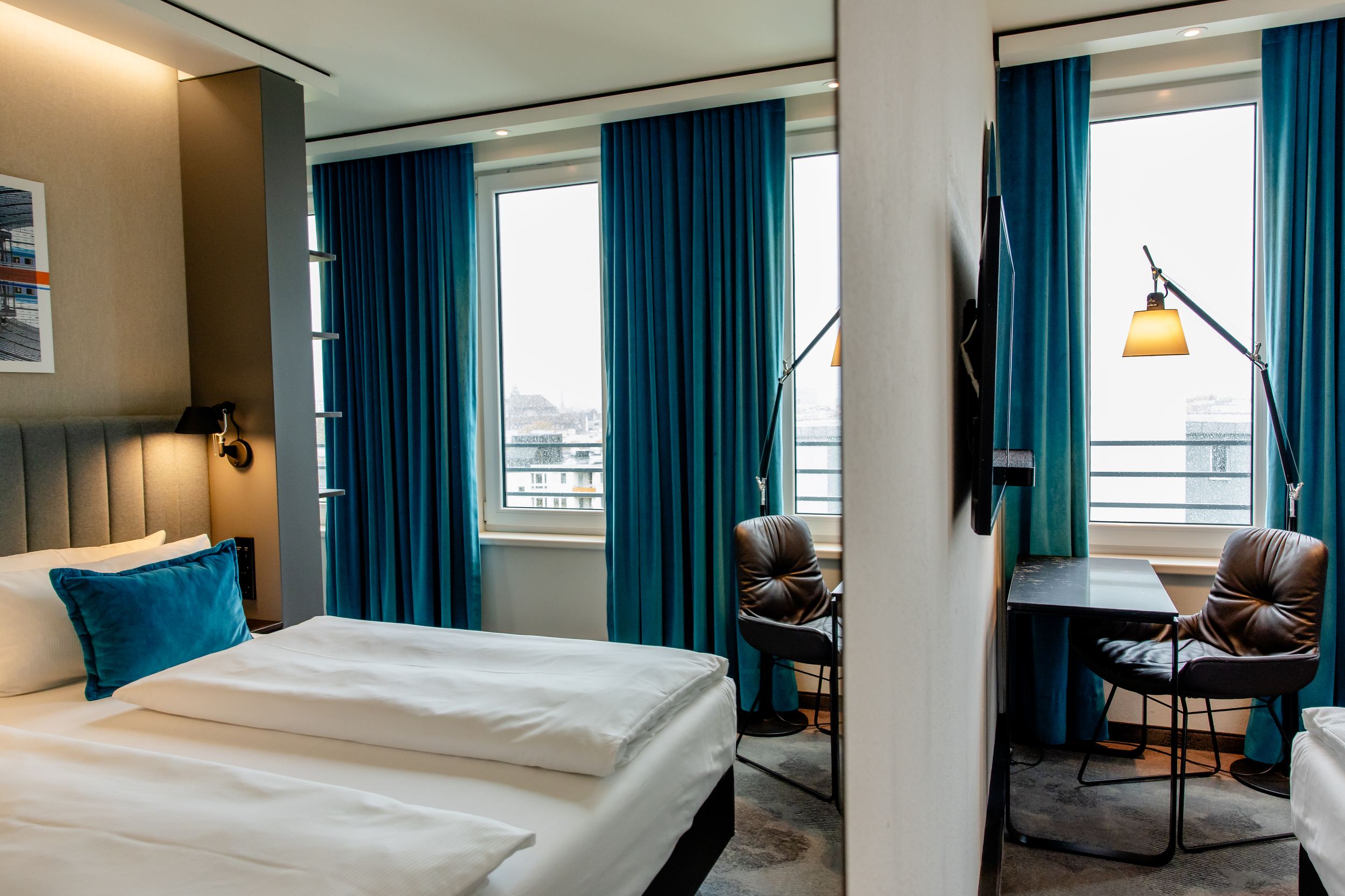 Hotel Motel One Berlin-Hauptbahnhof Modern eingerichtetes Hotelzimmer mit gemütlichem Doppelbett, großen Fenstern und blauem Vorhang. Arbeitsbereich mit Lederstuhl und Schreibtisch, angenehmes Licht und ruhige Atmosphäre für Erholung und produktives Arbeiten.