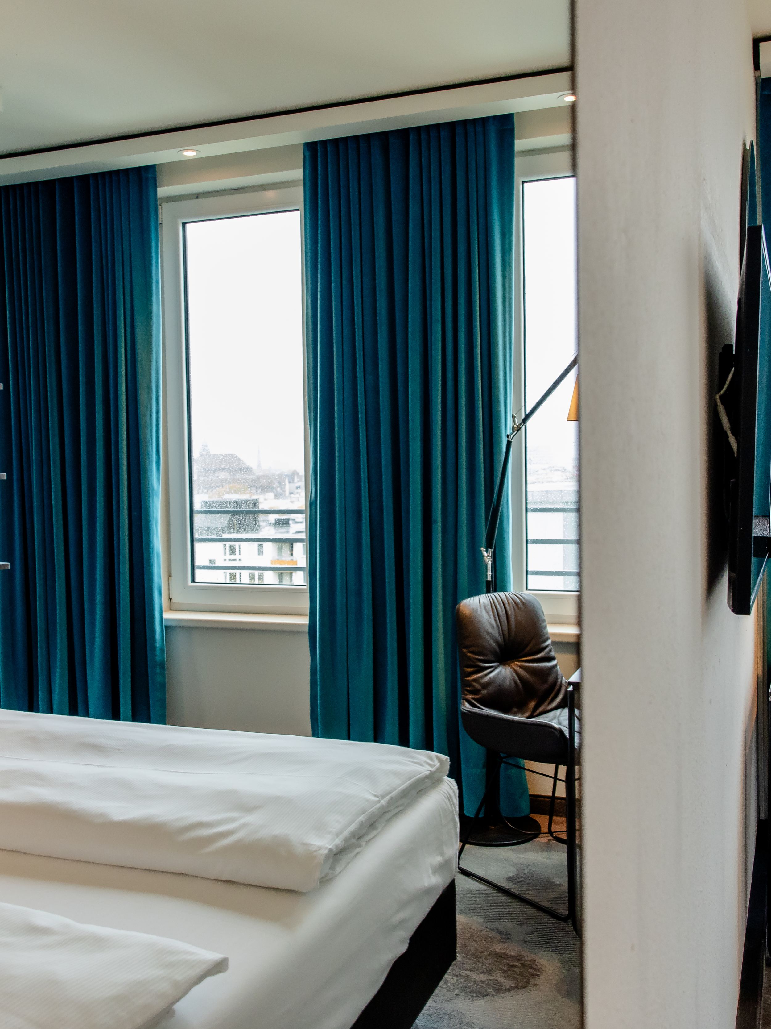 Hotel Motel One Berlin-Hauptbahnhof Modern eingerichtetes Hotelzimmer mit gemütlichem Doppelbett, großen Fenstern und blauem Vorhang. Arbeitsbereich mit Lederstuhl und Schreibtisch, angenehmes Licht und ruhige Atmosphäre für Erholung und produktives Arbeiten.