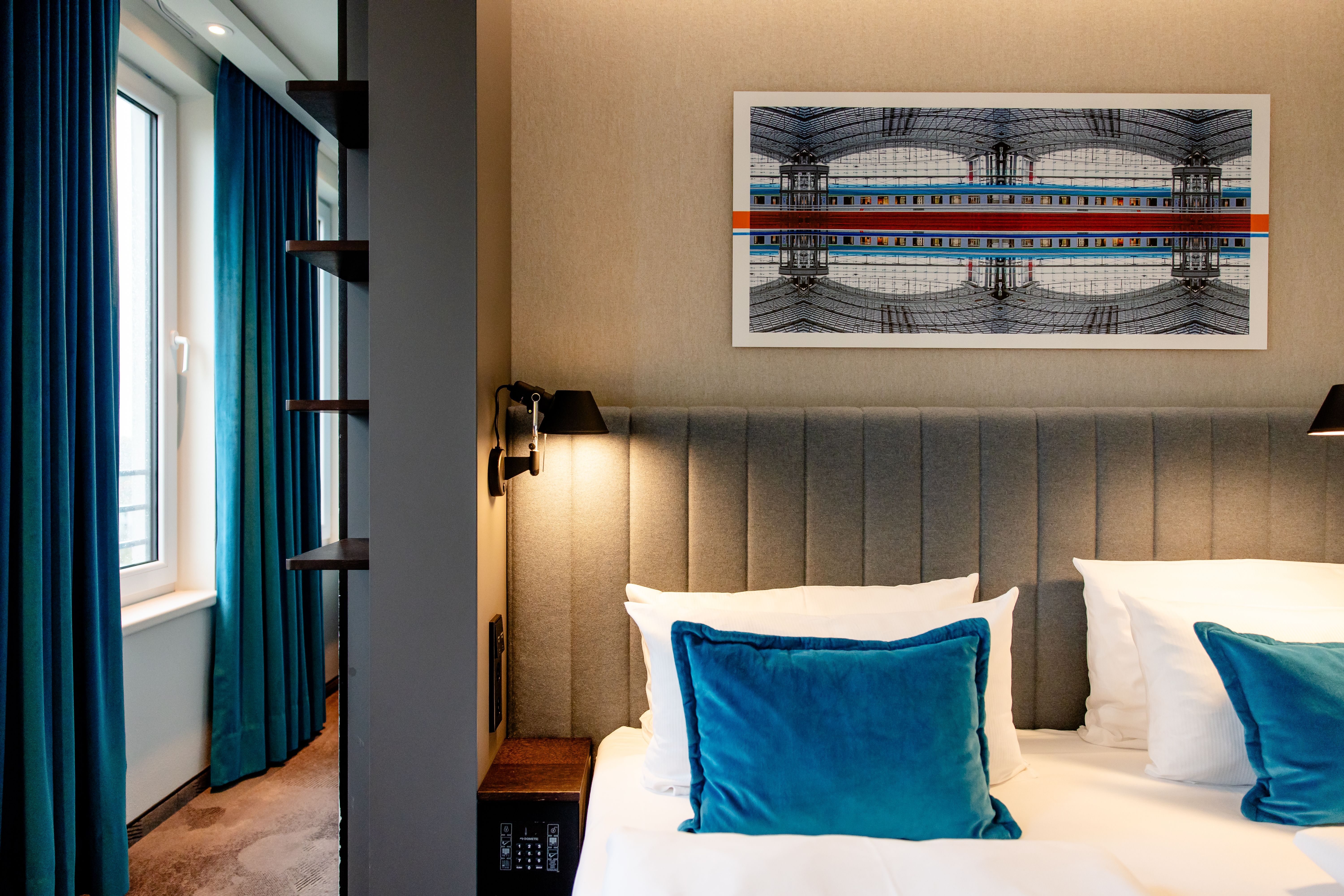 Hotel Motel One Berlin-Hauptbahnhof Helles Hotelzimmer mit bequemem Doppelbett, grauem gepolstertem Kopfteil und blauen Samtkissen. Neben dem Bett Wandlampen und großes Fenster mit blauen Vorhängen. Moderne, ruhige Atmosphäre, ideal zum Entspannen.
