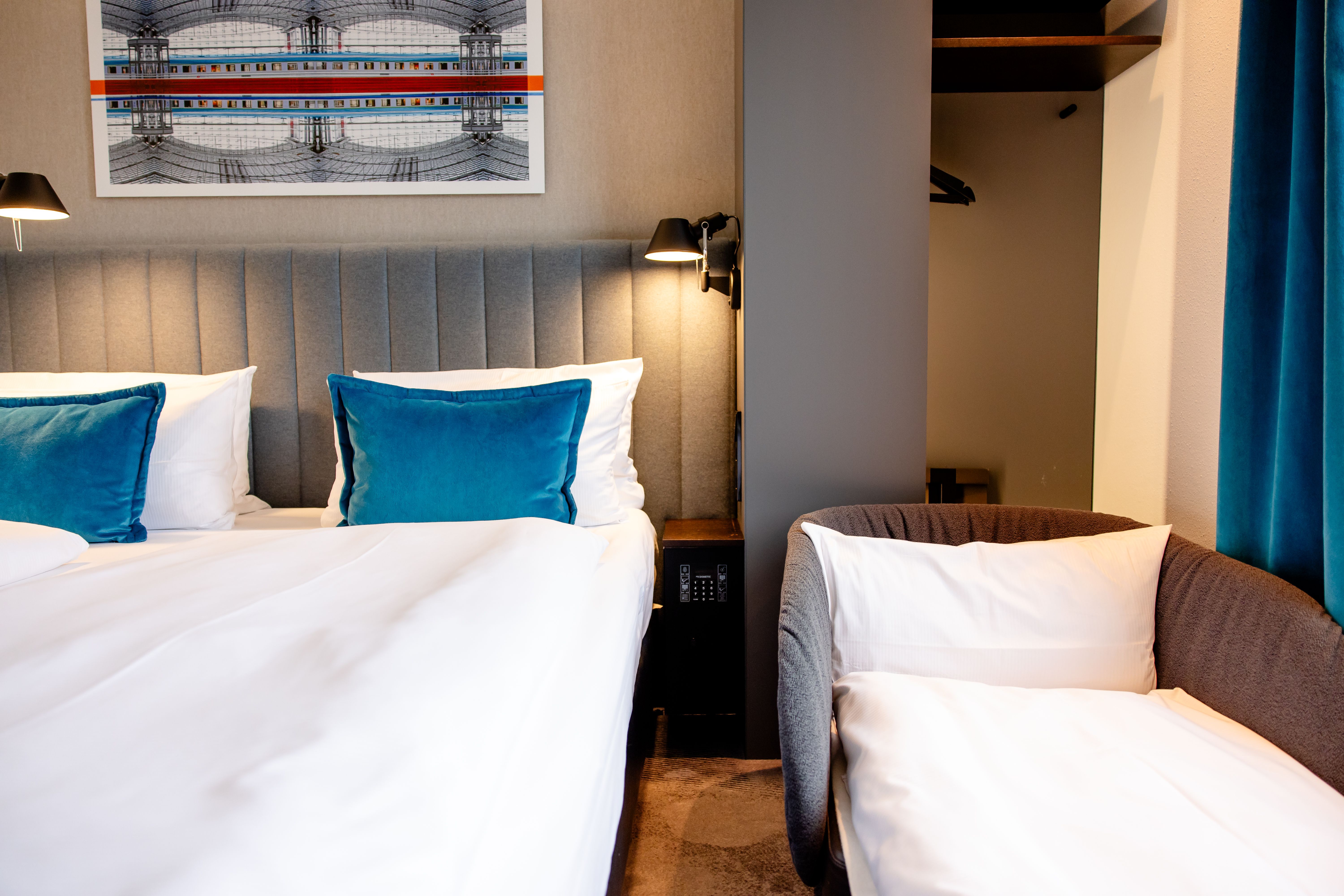 Hotel Motel One Berlin-Hauptbahnhof Modern eingerichtetes Hotelzimmer mit großem Bett, kräftig blauen Kissen, gemütlichem Sessel und offenem Kleiderschrank. Sanfte Beleuchtung schafft eine ruhige Atmosphäre.