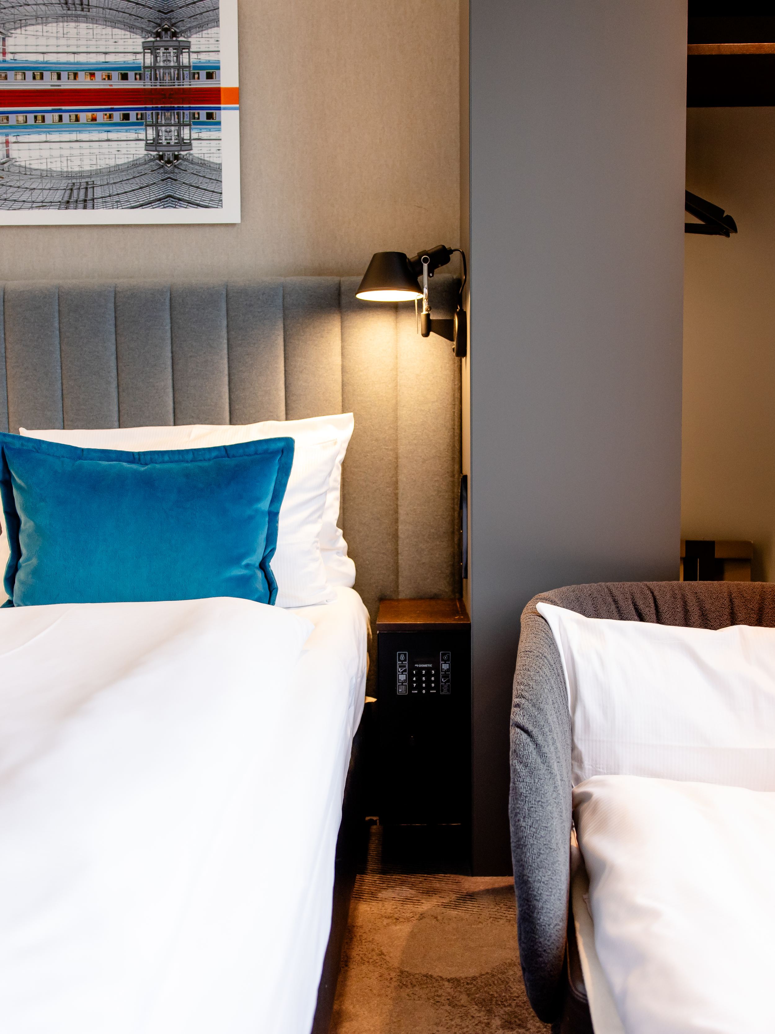 Hotel Motel One Berlin-Hauptbahnhof Modern eingerichtetes Hotelzimmer mit großem Bett, kräftig blauen Kissen, gemütlichem Sessel und offenem Kleiderschrank. Sanfte Beleuchtung schafft eine ruhige Atmosphäre.