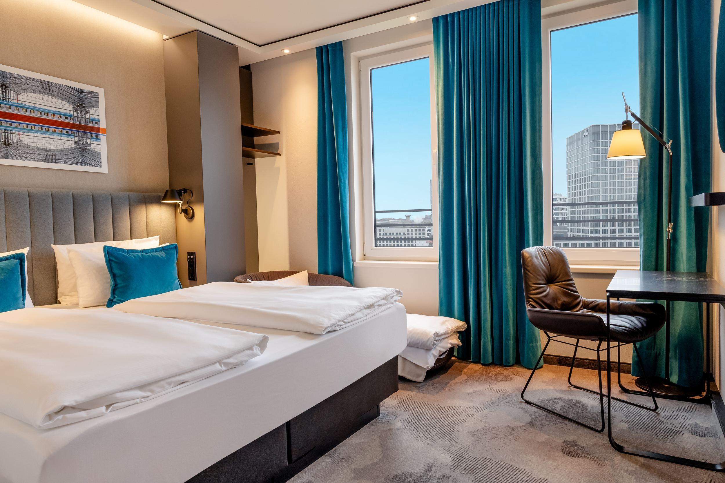 Hotel Motel One Berlin-Hauptbahnhof Modern eingerichtetes Hotelzimmer mit Doppelbett, blauen Vorhängen und großen Fenstern, die Stadtblick bieten. Schreibtisch mit Stuhl und Leselampe neben den Fenstern für komfortables Arbeiten oder Lesen. Helle, gemütliche Atmosphäre für erholsamen Aufenthalt.