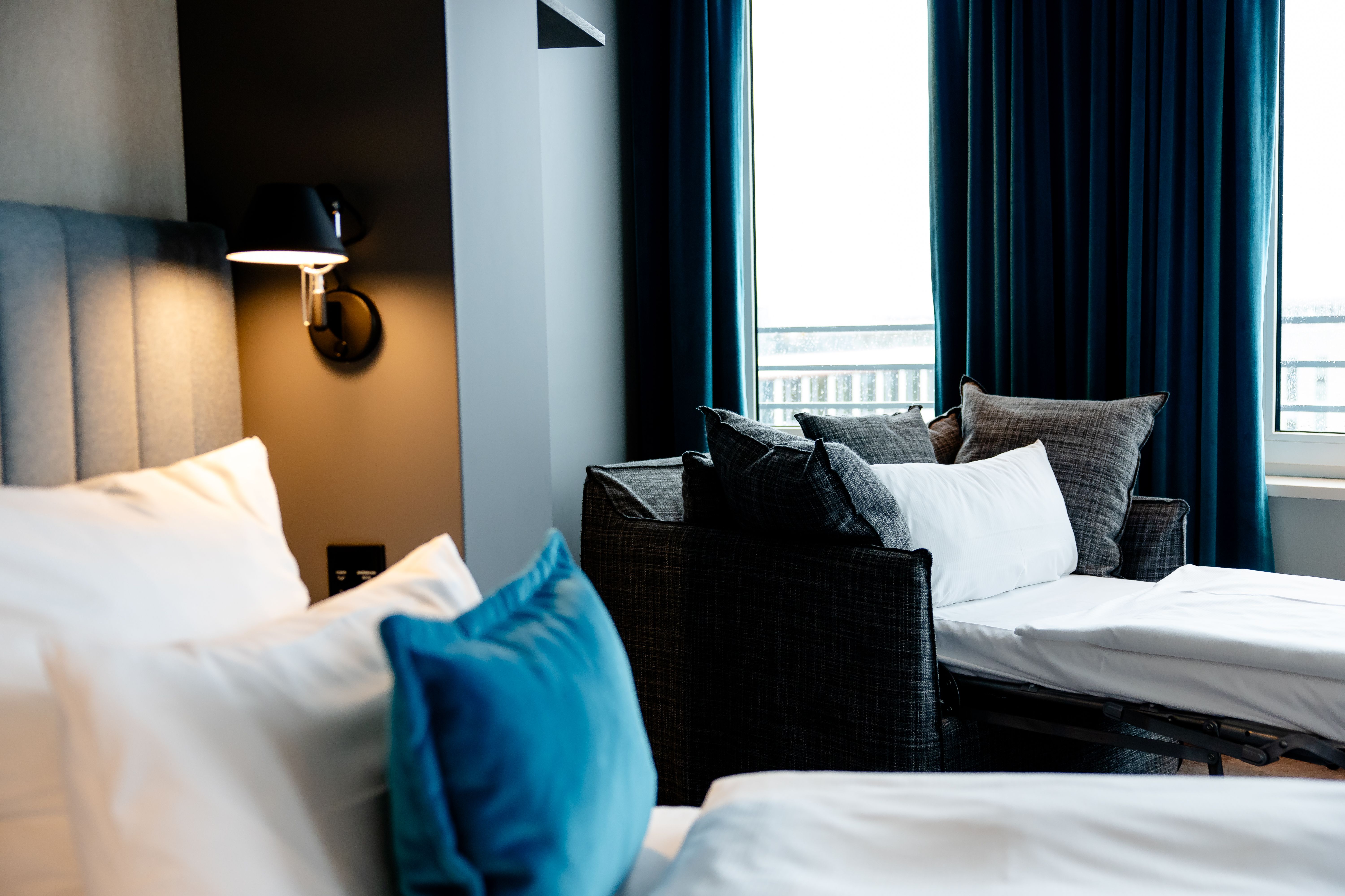 Hotel Motel One Berlin-Hauptbahnhof Gemütliches Hotelzimmer mit bequemen Betten und blau-weißen Kissen, indirekter Wandbeleuchtung und großen Fenstern mit dunklen Vorhängen, die viel Tageslicht und einen Ausblick bieten.