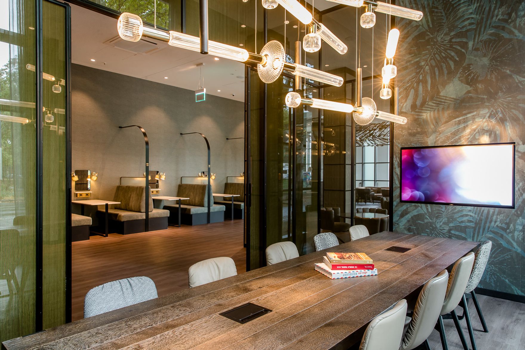 Hotel Berlin Hauptbahnhof Motel One heller Raum mit langem Tisch, mehreren Stühlen und Fernseher an der Wand sowie Frühstücksbereich im Hintergrund