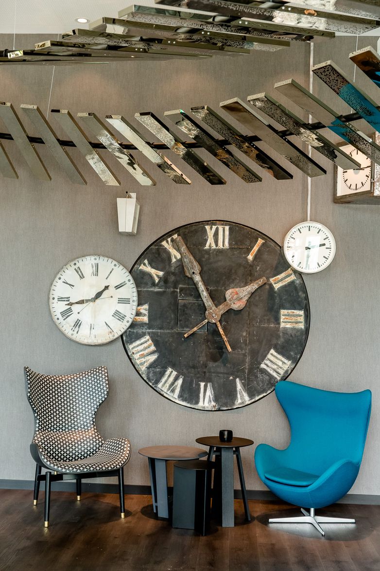 Designhotel Berlin Hauptbahnhof Motel One Blick auf einen Stuhl und einen Egg Chair mit Beistelltischen in deren Mitte sowie mehrere Uhren an der Wand