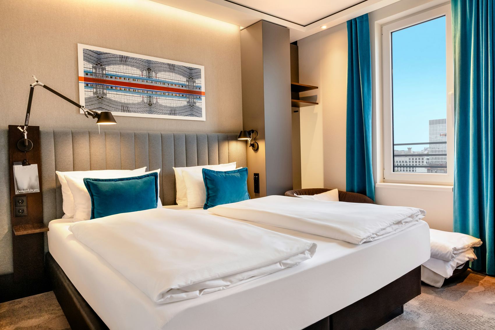 Hotel Motel One Berlin-Hauptbahnhof Modern eingerichtetes Hotelzimmer mit großem Doppelbett, hellen Wänden, blauen Akzentkissen und Vorhängen sowie großem Fenster mit Stadtblick.