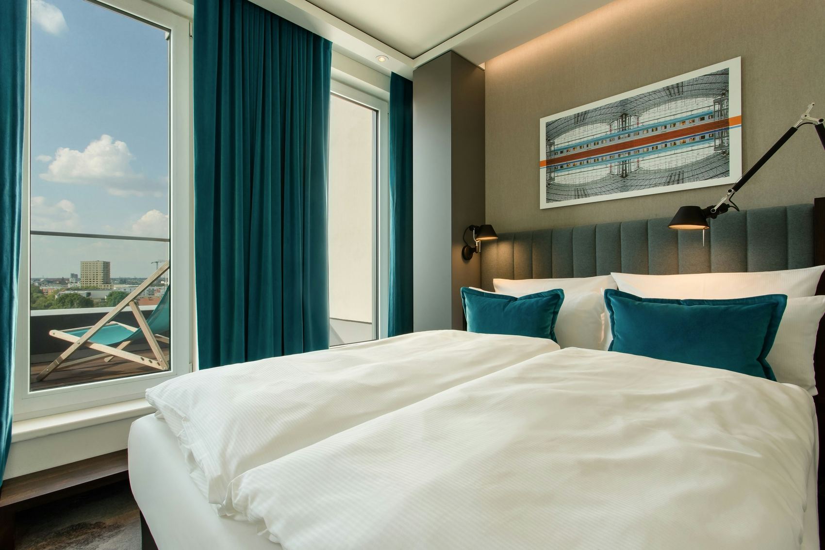 Hotel Motel One Berlin-Hauptbahnhof Modern eingerichtetes Hotelzimmer mit großem, weiß bezogenem Doppelbett, türkisfarbenen Kissen und Vorhängen. Große Fenster bieten Blick auf einen Balkon mit Liegestuhl und die Stadt im Hintergrund. Ruhige, einladende Atmosphäre für erholsamen Aufenthalt.