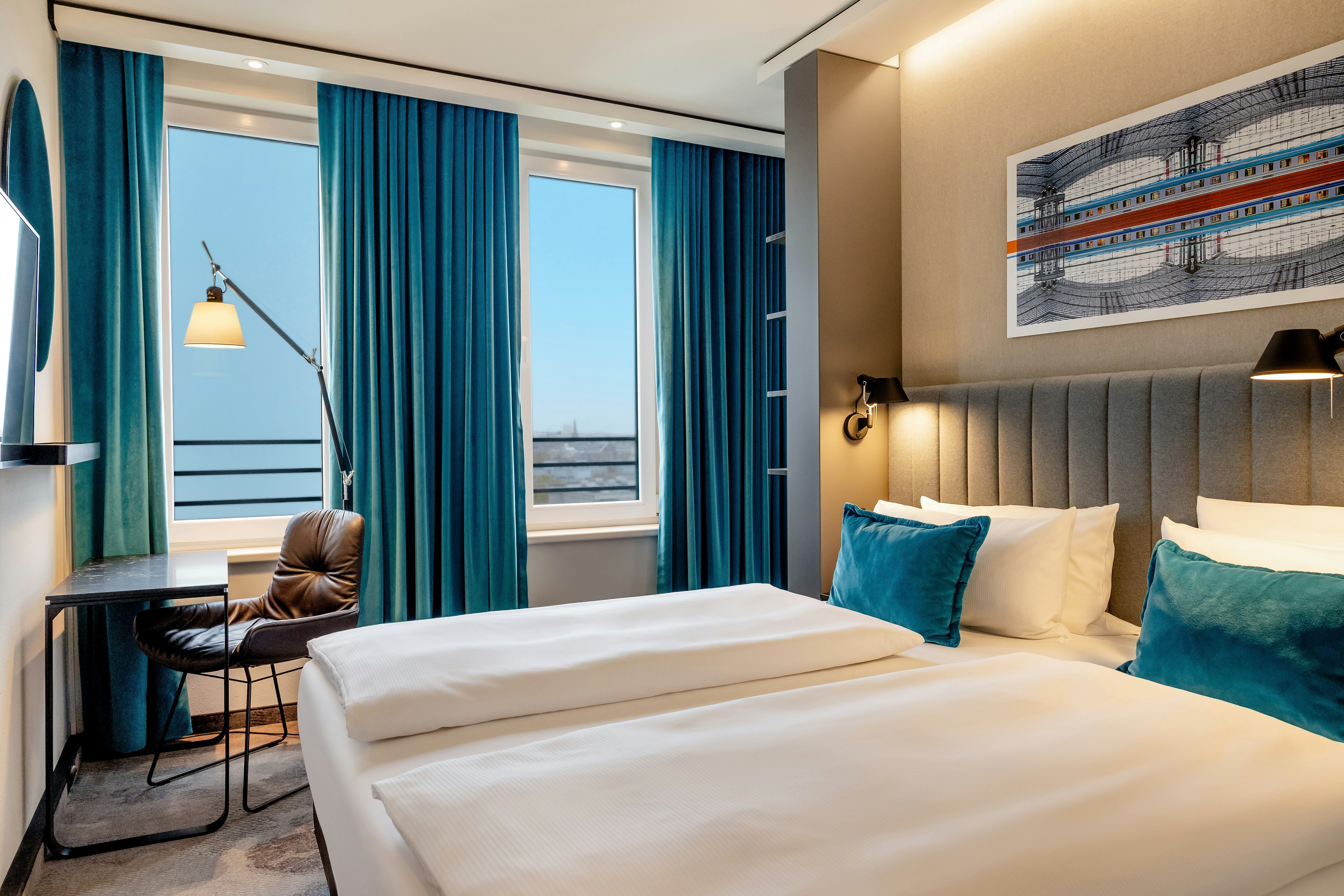 Hotel Motel One Berlin-Hauptbahnhof Modernes Hotelzimmer mit zwei Einzelbetten, blauen Akzenten, großem Fenster mit Blick ins Freie und einem komfortablen Arbeitsbereich. Helle, freundliche Atmosphäre für Erholung und produktives Arbeiten.